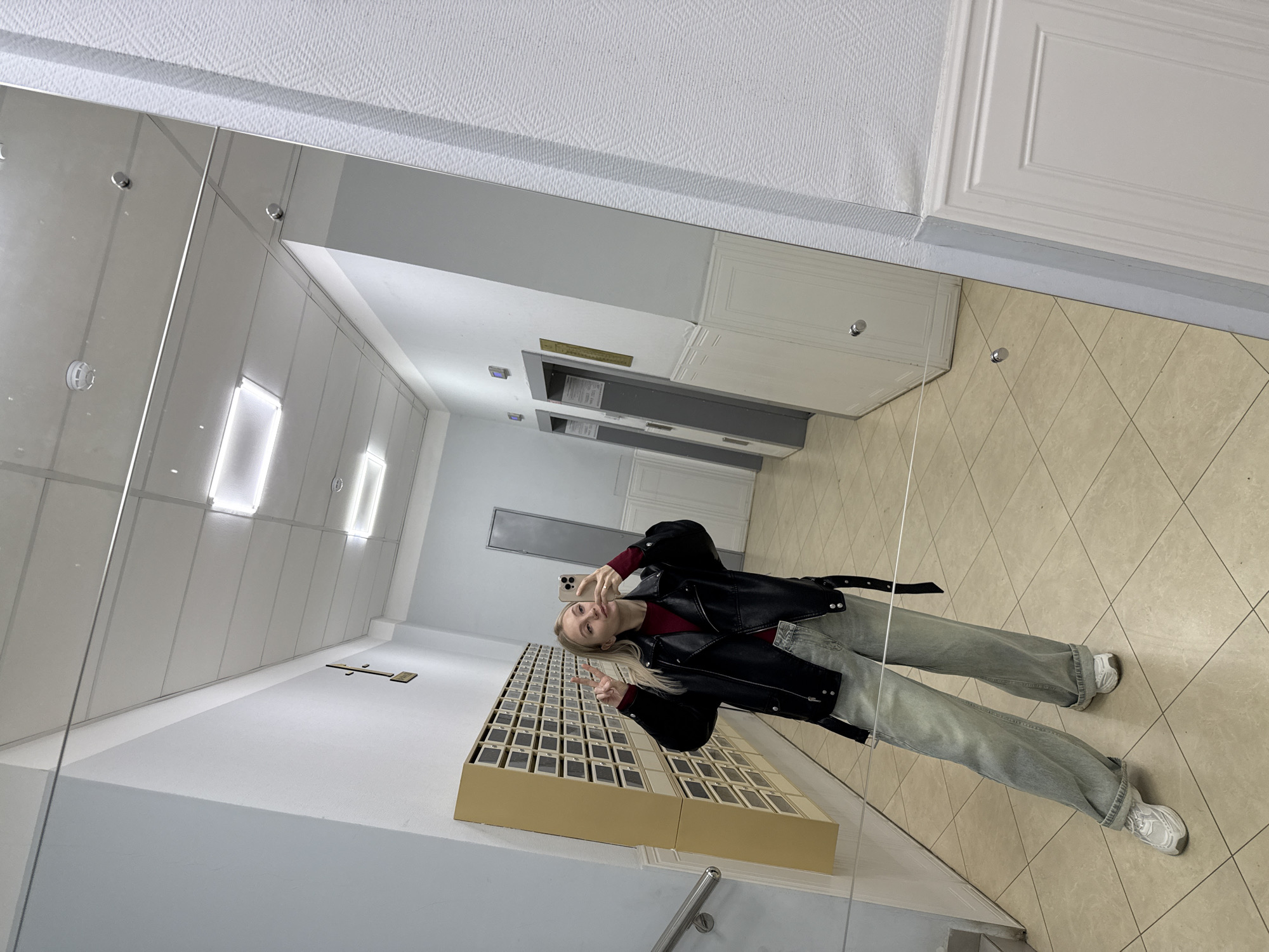 A woman stands in the reflection of a mirrored wall, holding a smartphone and making a peace sign. She is wearing a black leather jacket, a maroon top, light blue jeans, and white sneakers. Behind her is a wall with multiple mailboxes. The background includes a tiled floor, white walls, and fluorescent lighting. The location is Izhevsk, Russia.
