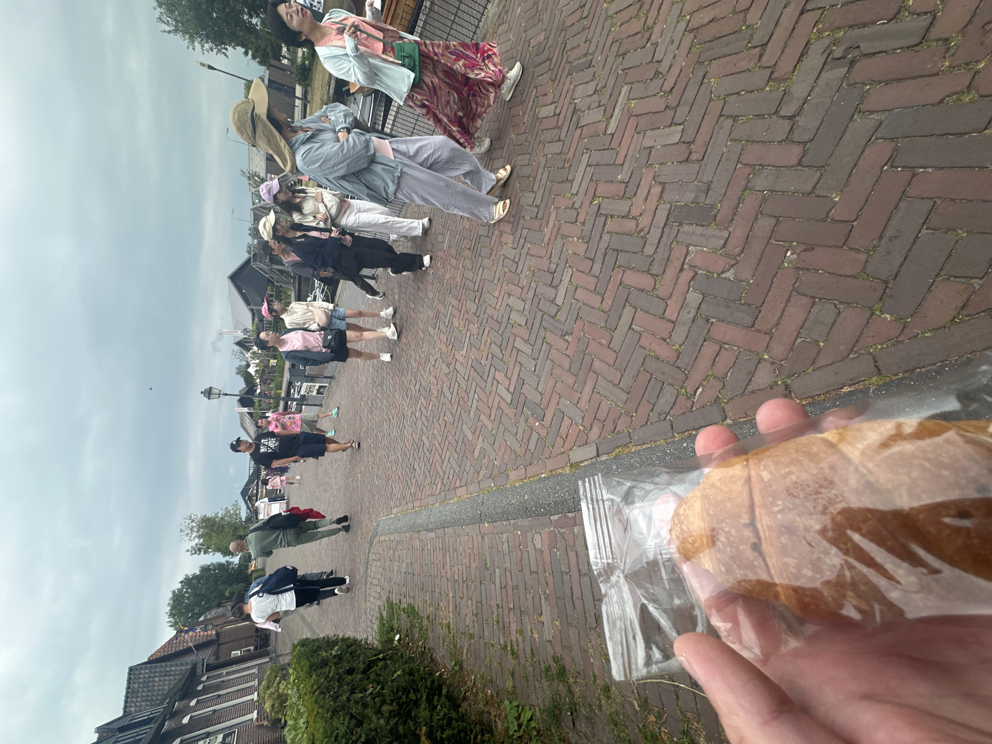 A person's hand holding a pastry in a clear plastic bag is in the foreground, obscuring the bottom of the image. Behind the hand, a brick-paved pathway, laid in a herringbone pattern, is visible. Several people are walking on the pathway, some in groups, heading away from the viewer. They are dressed in casual, warm-weather clothing. To the left of the pathway, a row of traditional Dutch houses with dark tiled roofs and brick facades is visible, partially obscured by lush green hedges. Beyond the pathway and houses, a cloudy sky looms overhead. The scene appears to be in Giethoorn, Netherlands, a village known for its canals and lack of roads, where pathways are common.