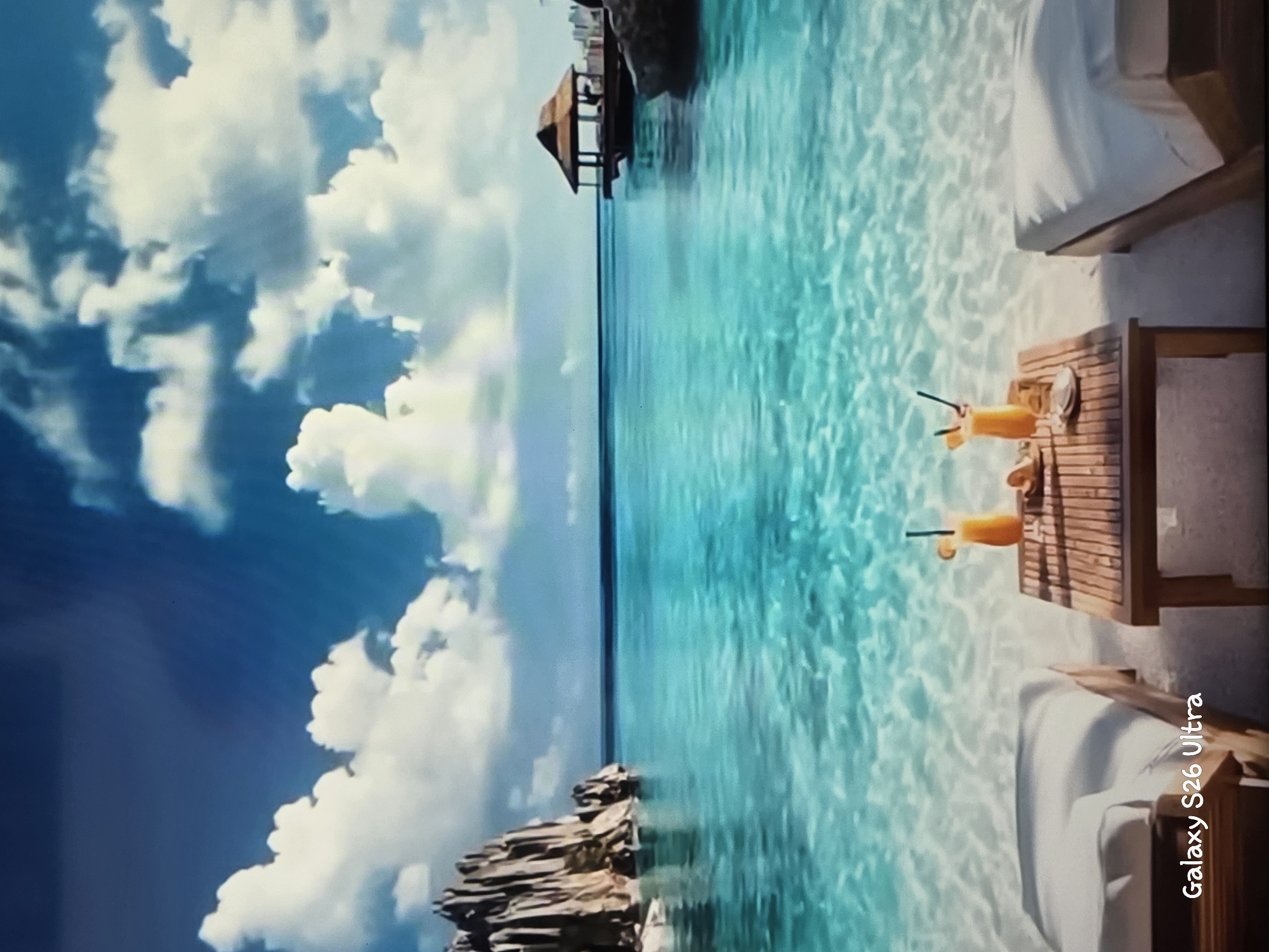A picturesque tropical beach scene unfolds, featuring crystal-clear turquoise water with sunlight illuminating the sandy bottom. In the foreground, two lounge chairs are set up on a wooden deck next to a small table. On the table, there are two glasses of orange-colored drinks with straws, suggesting a relaxing moment. Beyond the chairs, the water stretches out to meet a clear blue sky filled with fluffy white clouds. A small wooden gazebo or hut is visible in the distance, perched over the water, adding to the serene and secluded atmosphere. To the left of the frame, a rocky outcrop juts out into the water, contrasting with the smooth sand and calm sea. The lighting suggests it is daytime, possibly late morning or early afternoon, with bright sunshine. The overall mood is tranquil and inviting, evoking a sense of a peaceful vacation. The visible text reads "Galaxy S26 Ultra" in white lettering.
