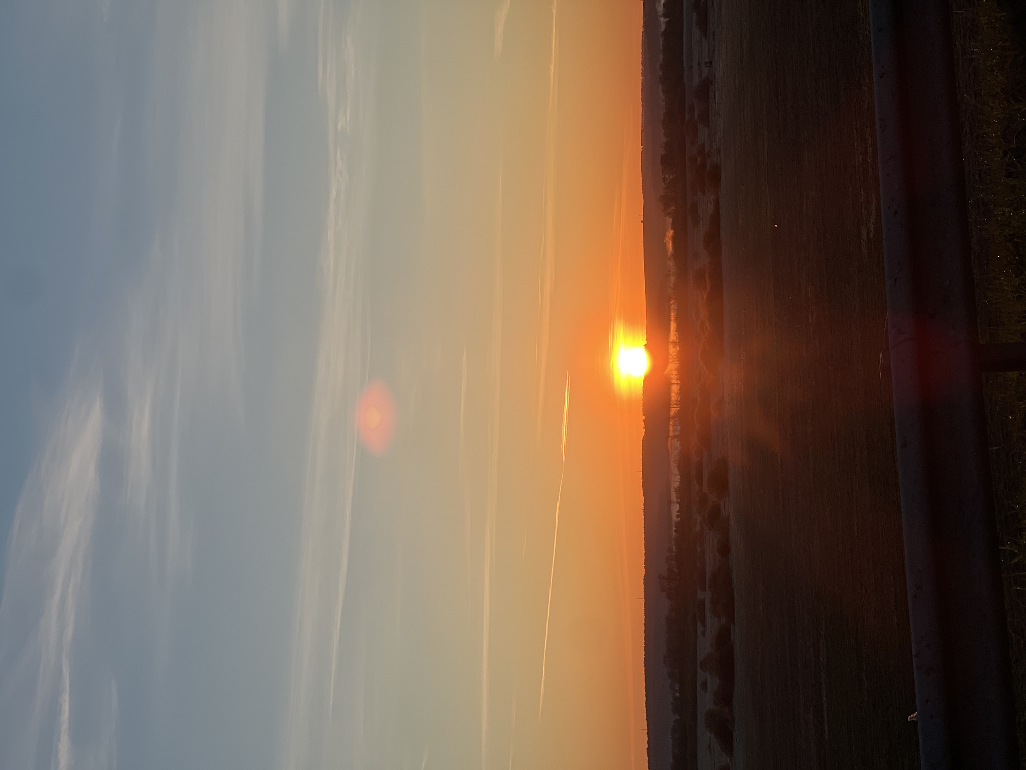 A sunset is occurring near Izhevsk, Russia. The sun is visible near the horizon, shining brightly with orange and yellow hues. The sky transitions from blue to orange. A field with a treeline sits on the right side of the frame, alongside a pole. Jet contrails cross the sky.