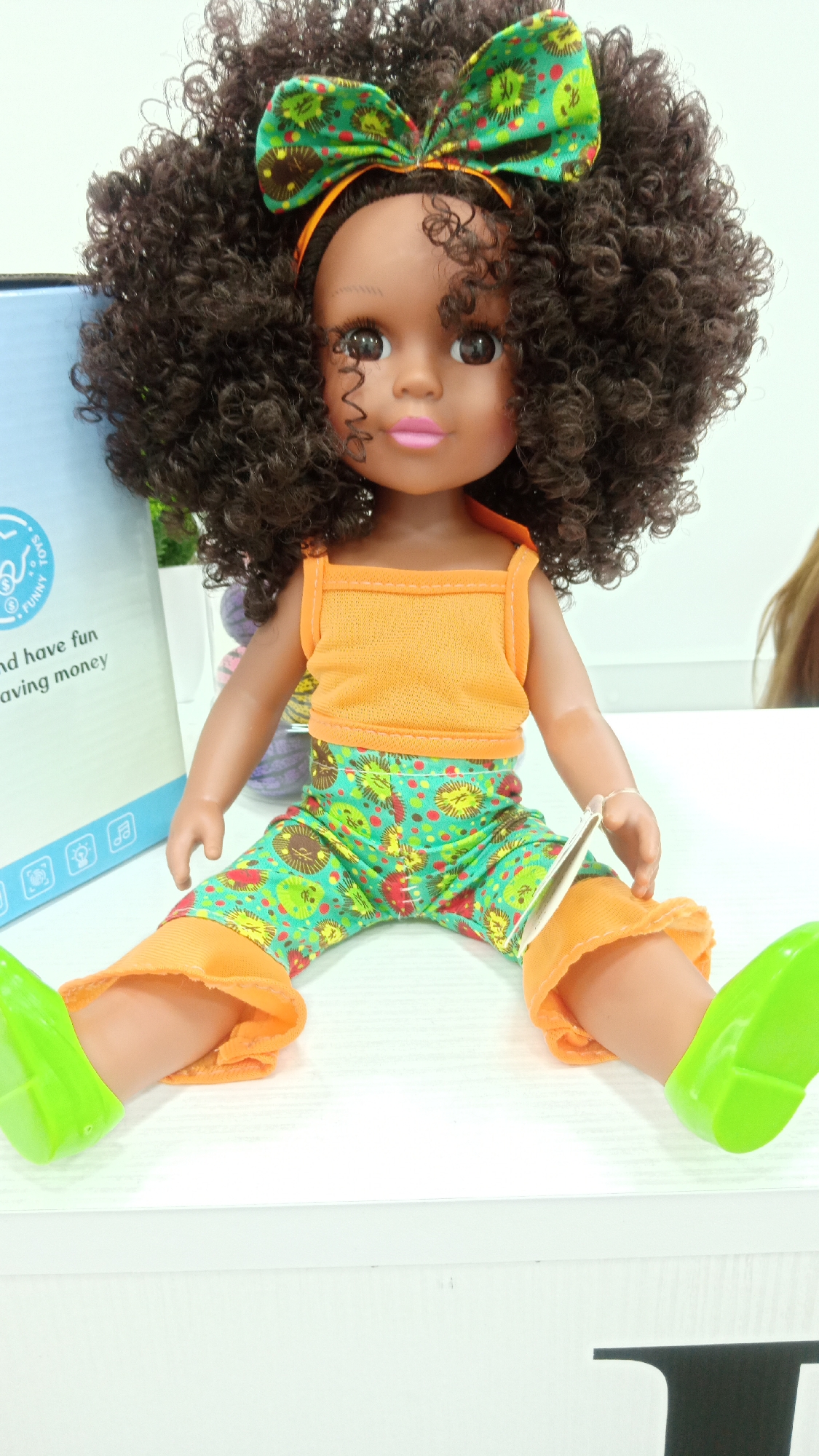 A doll with dark brown curly hair and an orange headband with a green patterned bow is sitting on a white surface. The doll has brown skin, pink lips, and brown eyes. It is wearing an orange tank top and green patterned pants with orange ruffles at the bottom. The doll has green plastic shoes. A box with the words "and have fun saving money" can be seen in the background on the left. The environment is indoors. The location is not identifiable as being Yerevan, Armenia.