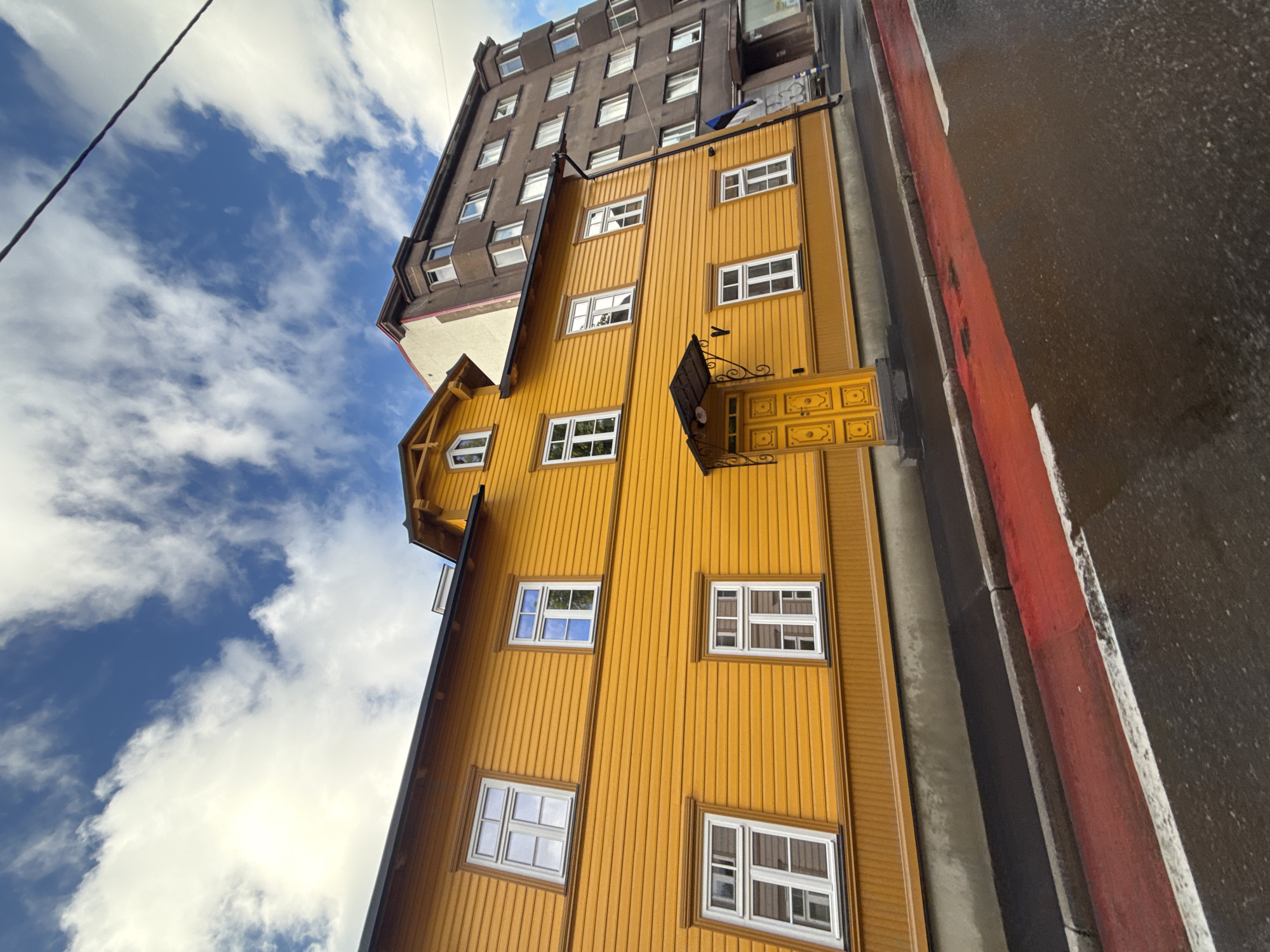 The image depicts a narrow, vertically oriented view of a yellow wooden building next to a taller, darker building, under a cloudy sky. The yellow building has a facade of vertical wooden planks and several white-framed windows, some with multiple panes. A decorative yellow door with carved panels is visible on the second story, with an ornate metal sign hanging above it. A narrow, dark asphalt road with a red and white painted curb runs alongside the buildings. The sky is a mix of blue and white clouds, suggesting daylight. The perspective is angled upwards, emphasizing the height of the buildings. The scene appears to be urban, with architectural elements suggesting a European setting, consistent with the provided location context of Tallinn, Estonia. There is no visible text in the image.