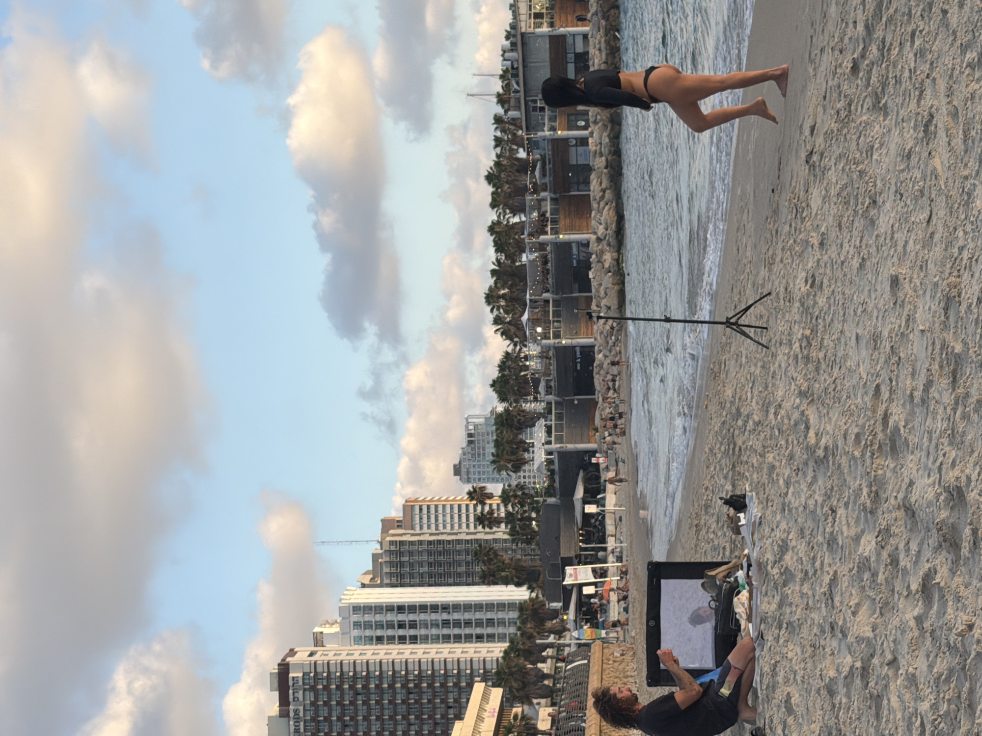 The image depicts a beach scene in Tel Aviv, Israel, with the sky filling the left side of the frame. The sky is a pale blue, dotted with fluffy, light-colored clouds, suggesting it is either early morning or late afternoon. The beach itself is sandy, with waves gently lapping at the shore on the right.
In the foreground, a woman is positioned on the sand, leaning forward with her hands on the ground, as if in a plank or push-up pose. She is wearing a black top and bikini bottoms. Further to the right and closer to the water, a man is seated on the sand, facing away from the viewer and towards a portable screen or easel. He has curly brown hair and is wearing a dark shirt and blue shorts.
Behind the beach, a row of buildings and palm trees can be seen. The buildings are modern, multi-story structures with many windows, characteristic of a coastal city. A structure with a dark roof and wooden accents runs along the beachfront, possibly housing shops or cafes. The overall atmosphere appears relaxed, with people enjoying the beach. The presence of a tripod suggests that either the woman or the man is involved in some form of photography or videography. The lighting indicates it is not the peak of midday.