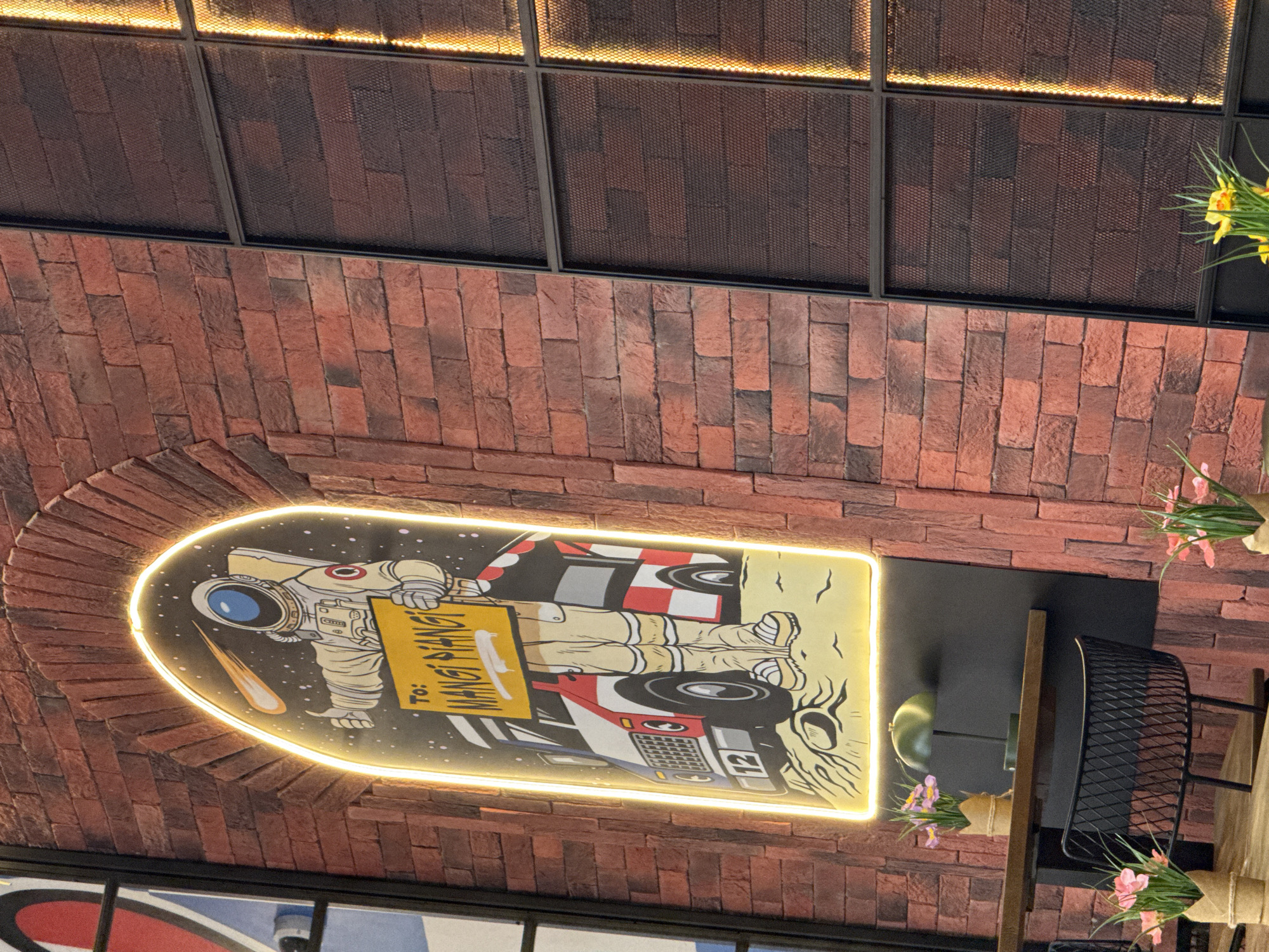 The image depicts an interior setting with a visible ceiling featuring a grid-like structure illuminated from above. A brick wall is prominently displayed, featuring a rounded archway where a brightly lit sign or mural is mounted. The sign depicts an astronaut holding a sign that reads "To: Mangi Pang". Adjacent to the sign, there's a section of what appears to be a decorative installation consisting of a black metal basket mounted on a wooden structure, with floral arrangements nearby. Based on the text on the sign, this could be a restaurant or business located in Sofia, Bulgaria named Mangi Pang.