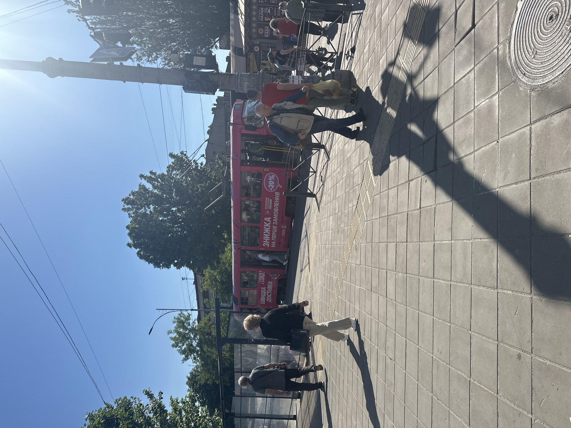 The image captures a sunny urban street scene featuring a red tram (or trolleybus) alongside a sidewalk where several people are present. In the foreground, long shadows stretch across the grey square-tiled pavement, indicating bright sunlight. A circular manhole cover is visible on the sidewalk.
Various individuals are depicted: a person in a light vest and dark pants walks near a metal barrier separating the sidewalk from the tram tracks, while another individual in a red shirt stands by the barrier, holding a yellow bag. Further along the sidewalk, a woman with light hair wearing a dark jacket and light pants walks away from the camera, followed by a man in a dark shirt and pants. Other figures are visible in the background and inside the tram.
The red tram is adorned with advertisements. Overhead wires are strung across the clear blue sky, with some tree foliage visible on either side. A street pole with a traffic light and a partially obscured sign is on the left. Buildings with storefronts can be seen in the background behind the tram stop.
The time of day appears to be sunny with a clear sky, evidenced by the bright light and distinct shadows. The overall setting is a bustling city environment during daytime.
Visible text on the tram includes "ШВИДКА LOKO! ДОСТАВКА" (SWIFT LOKO! DELIVERY) and an advertisement stating "ВСТАНОВЛЮЙ ЗНИЖКА ОСУНОК (-20%) НА ПЕРШЕ ЗАМОВЛЕННЯ" (INSTALL OSUNOK DISCOUNT (-20%) ON THE FIRST ORDER), likely promoting an app or service. The tram also bears the number "3324". In the background, shop signs display "artMobi," "НАПОЇ" (DRINKS), "KABA" (COFFEE), and "LATTE."