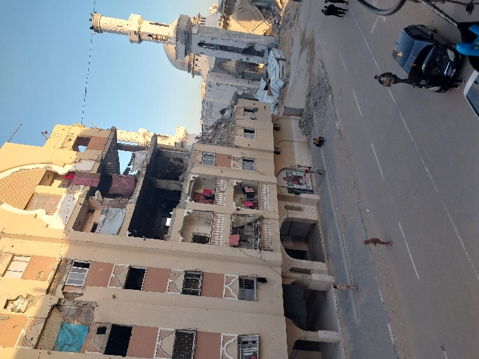 The image depicts a scene of extensive urban destruction, likely the aftermath of conflict or a major disaster. The overall setting is an outdoor street in what appears to be a Middle Eastern or North African city, given the architectural style including a prominent mosque.
In the foreground and mid-ground, on the left side, stands a large, multi-story building that has sustained severe damage. Large sections of its facade are missing, exposing the building's interior rooms and structural components. Charred areas and debris are visible within the exposed sections, suggesting fire damage. Personal items, including clothes, are seen hanging from what were once balconies or windows, indicating that the building was inhabited. The building's exterior, where intact, is a light, sandy beige color with some decorative patterns.
Further down the street, in the background, a white mosque with a tall minaret and a dome is visible. The minaret appears largely intact, though some wear or minor damage might be present at its pinnacle. The mosque structure itself, particularly its lower sections, also shows signs of damage, with rubble scattered around its base.
An asphalt road runs across the lower portion of the image. It has clear white lane markings. On the right side of the road, a dark-colored sedan, possibly black, is parked or stationary. Adjacent to the road and surrounding the damaged buildings, piles of rubble, concrete fragments, and other debris are scattered, further emphasizing the scale of the destruction.
Several people are visible in the scene, going about their daily activities amidst the ruin. A few individuals are standing near the parked car on the right. Other figures are walking or standing further down the street, closer to the mosque. One person is seen walking alone in the middle of the road in the foreground, and another is near the damaged building on the left. Their presence suggests an ongoing human presence and perhaps the initial stages of coping with or assessing the devastation.
The time of day appears to be mid-morning or early afternoon, indicated by the bright, clear blue sky and the distinct shadows cast by the buildings and people. The weather is fair and sunny. No discernible text is visible in the image. The scene conveys a powerful sense of loss and the profound impact of destructive events on urban environments and the lives of their inhabitants.