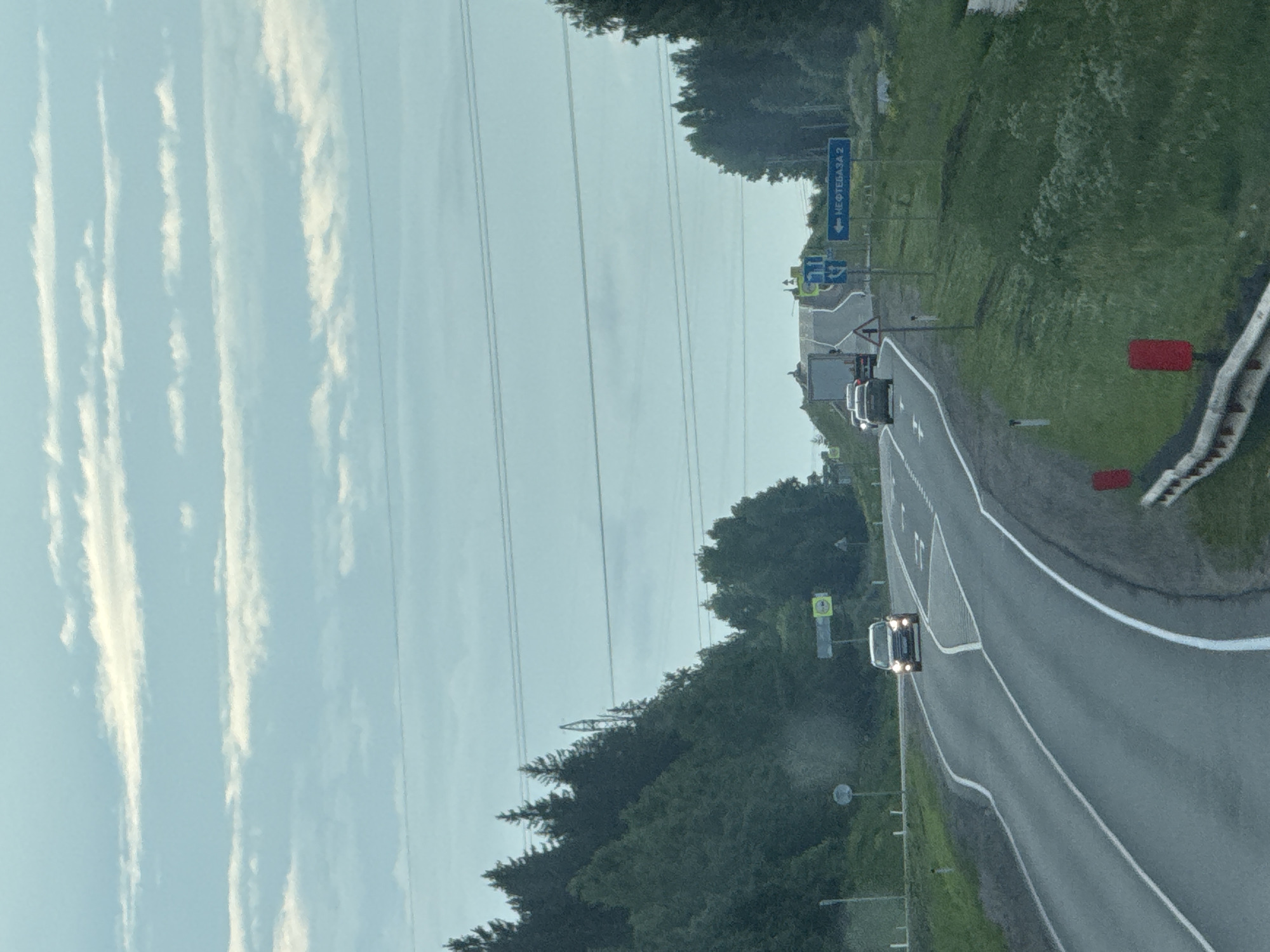 The image depicts a two-lane asphalt road curving through a forested landscape under a cloudy sky. A white SUV is approaching from the opposite direction, its headlights illuminated. A truck is visible further ahead on the same road, also traveling in the opposite direction. On the right side of the road, a blue directional sign points to the left, indicating "НЕФТЕБАЗА 2" (NefteBaza 2). The surrounding environment is characterized by dense green trees on both sides of the road, with a grassy embankment on the right. Power lines are strung across the upper portion of the frame. The scene suggests a rural or semi-rural setting.