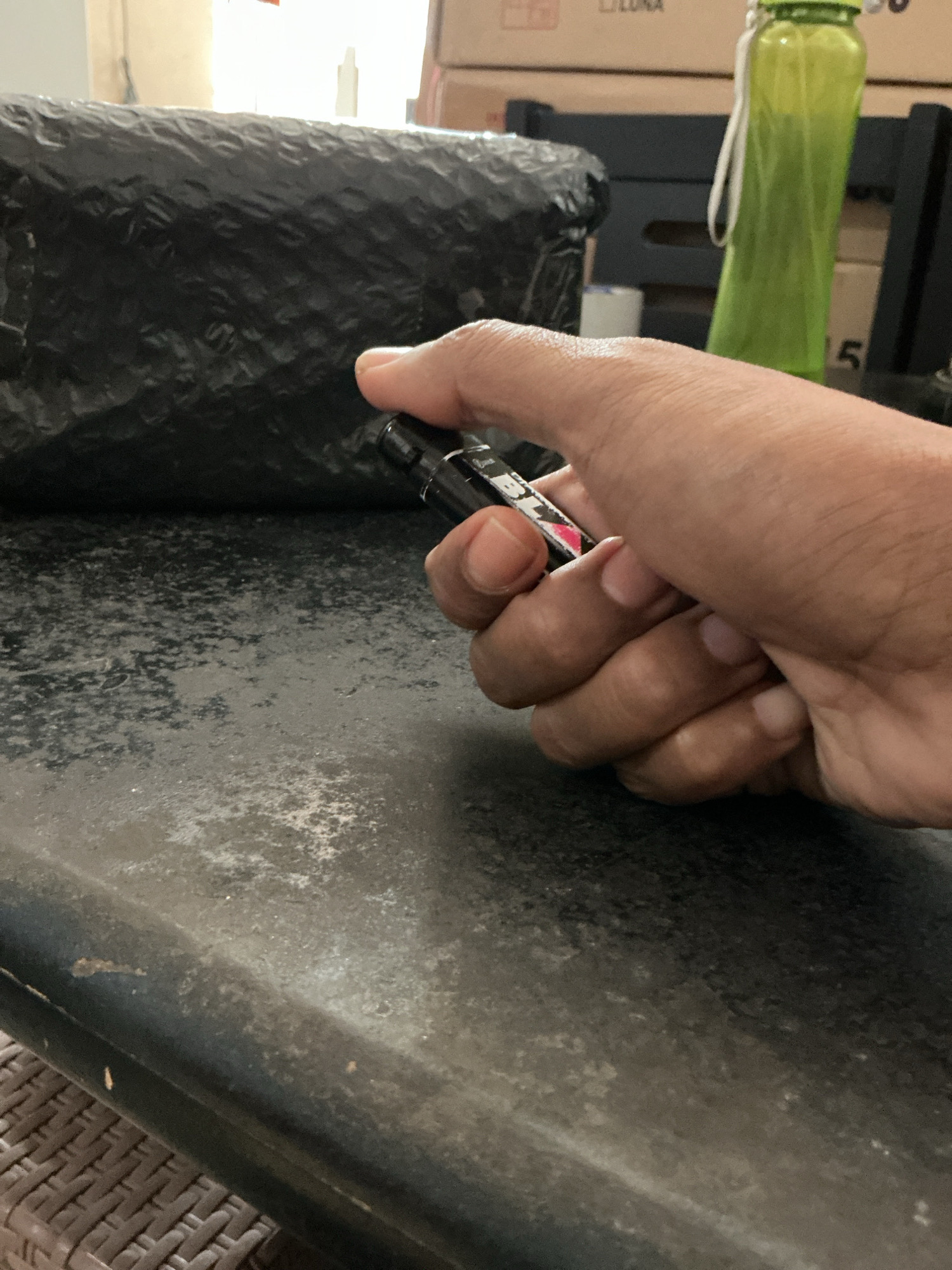 A hand is holding a black and silver object with a red accent. The object appears to be a pen or a similar small, handheld item. The background is blurred but suggests an indoor environment with boxes and a green water bottle visible. The surface in the foreground is dark and textured, possibly a table or countertop.
