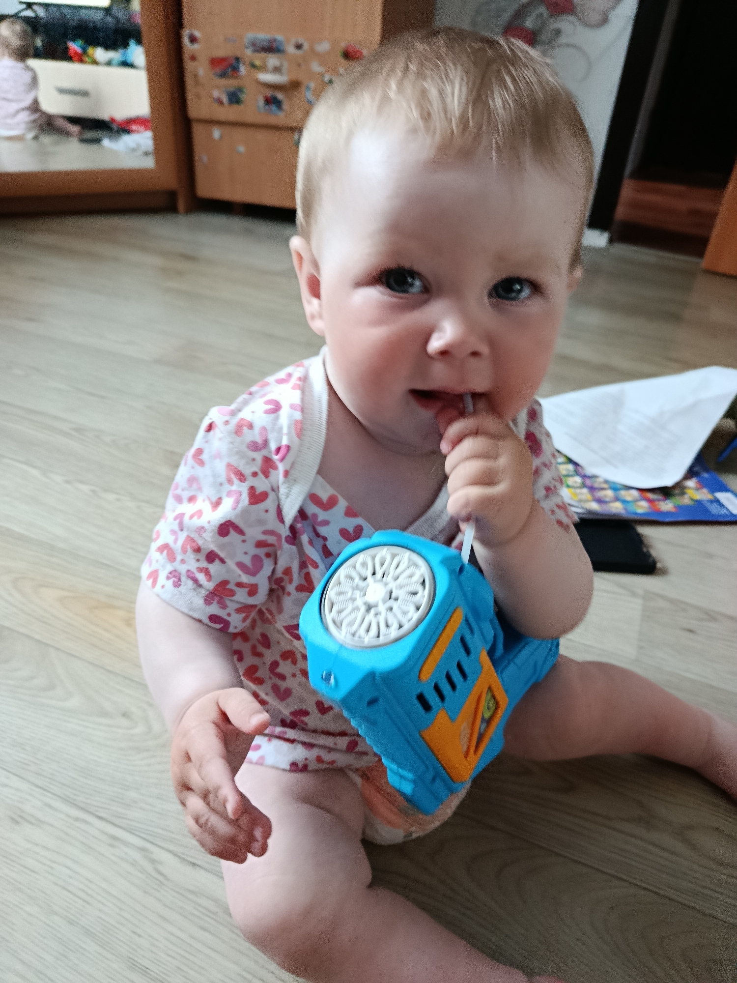 A baby with blond hair and blue eyes is sitting on a wooden floor. The baby is wearing a white onesie with small pink and red hearts. The baby is holding a blue and orange toy bubble gun in their right hand and has the wand of the bubble gun in their mouth. The background includes a wooden cabinet, a mirror reflecting a person in the room, and some papers and books on the floor. The location is Фоки, Russia.
