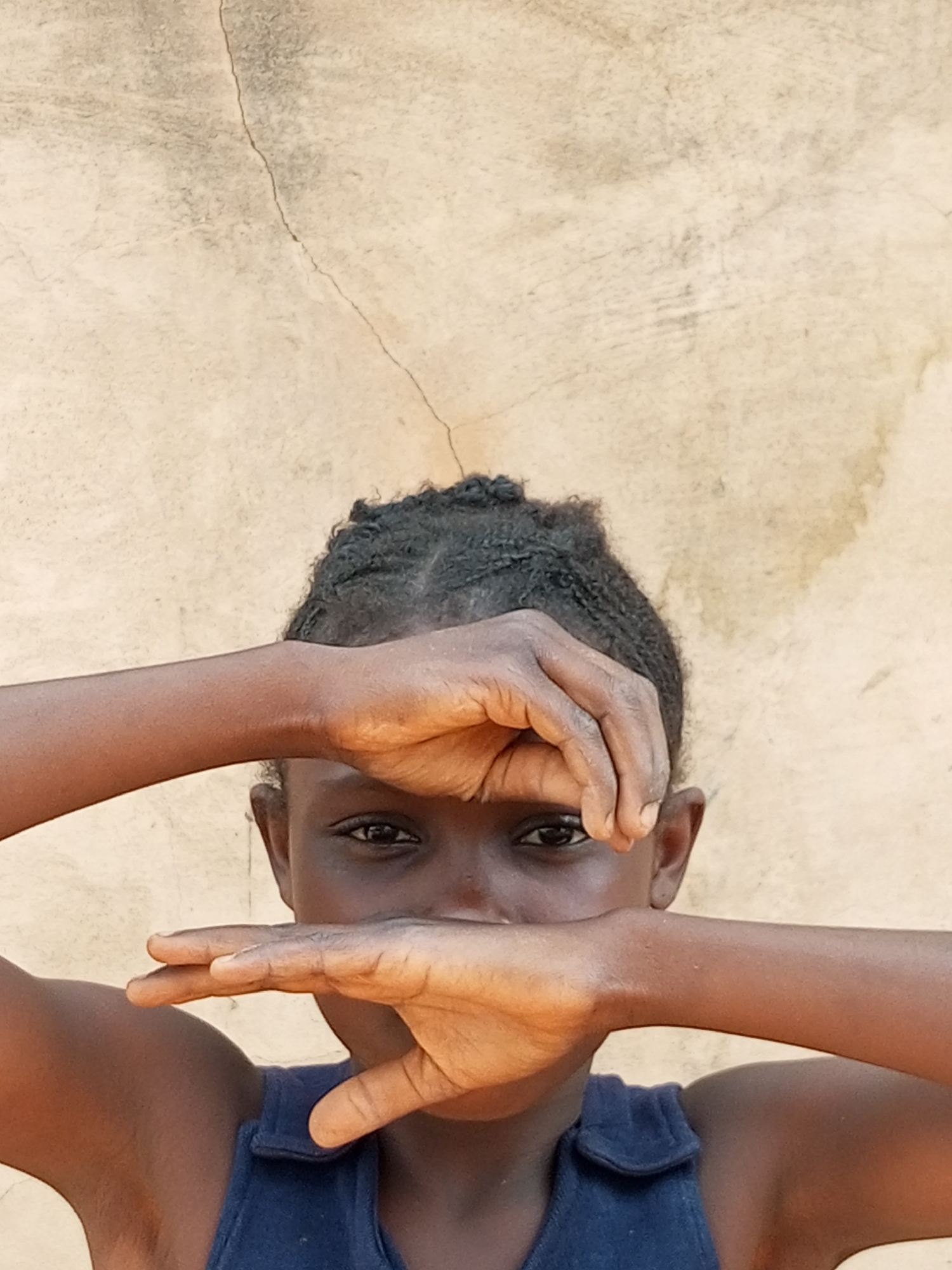 A young Black child is partially obscuring their face with their hands against a rough, light-colored wall with a crack running down the center. The child has dark, braided hair and is wearing a dark blue sleeveless garment. One eye is visible, looking directly at the camera. The lighting suggests an outdoor setting. The background wall appears weathered and textured, with some smudges and discoloration. The image is framed from the shoulders up, focusing on the child's face and hands.