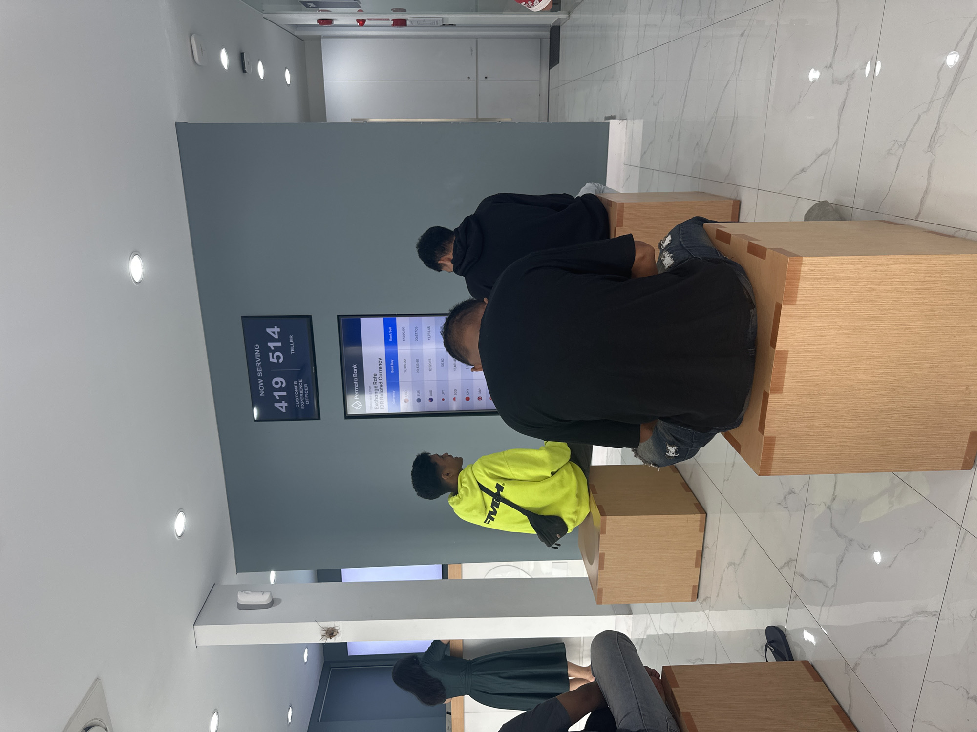 The image depicts an indoor waiting area, likely in a bank or service center, in Kuta Selatan, Indonesia. Several people are seated on wooden, box-like stools. A person wearing a bright yellow hoodie and a black shoulder bag is visible, along with other people wearing darker clothing. On the wall, digital displays indicate "Now Serving 419" and "514" for Customer Experience Officer and Teller, respectively. Another screen displays currency exchange rates for Permata Bank. The floor is tiled with white marble-like tiles. Overhead, recessed lighting is visible.