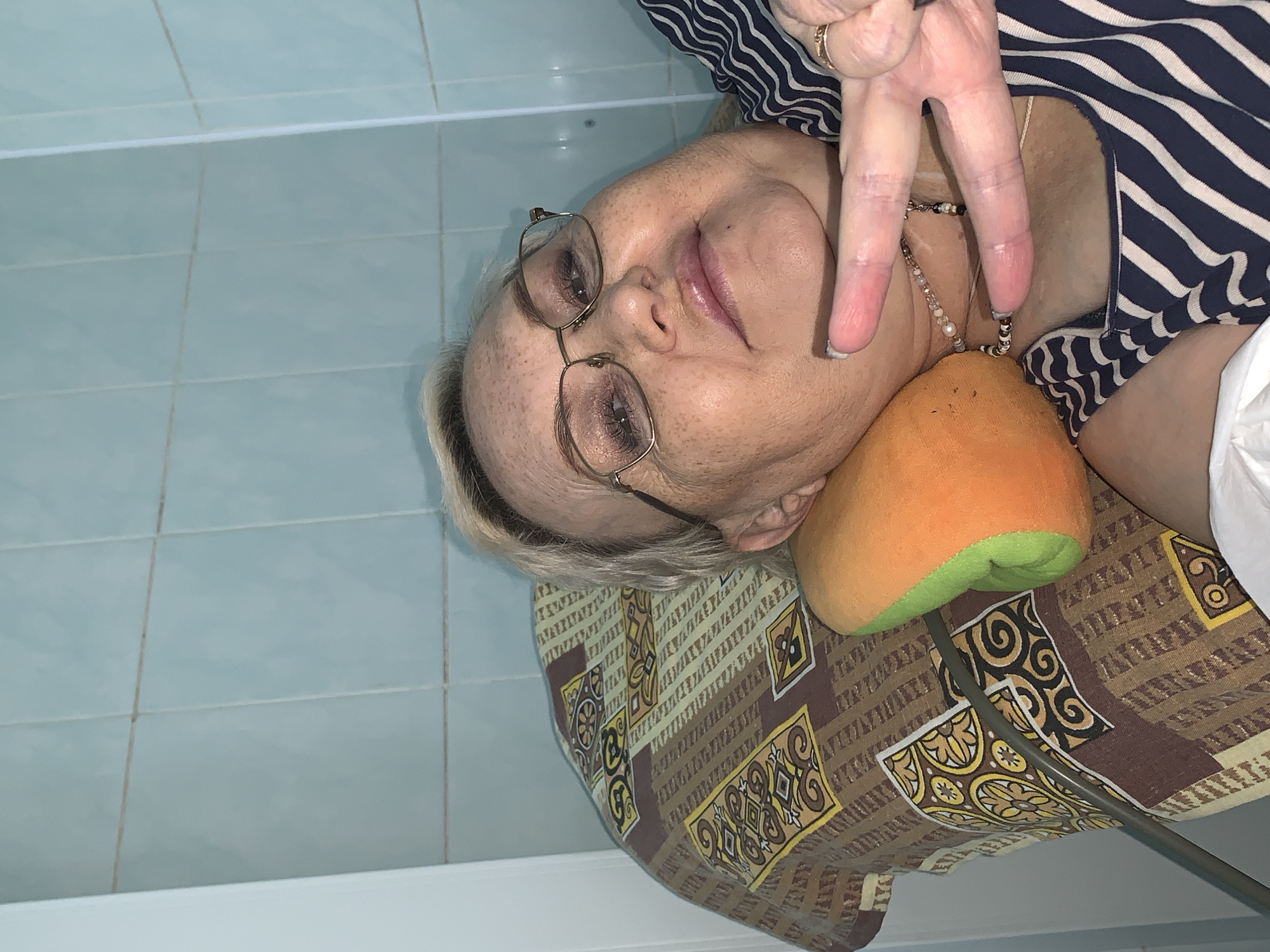 An elderly woman with glasses is lying on a patterned pillow, holding up her fingers in a peace sign. She is wearing a striped shirt and a necklace. The background is a tiled wall and a bathtub. It is not possible to determine the specific location in Chaikovsky, Russia, based on the provided image.