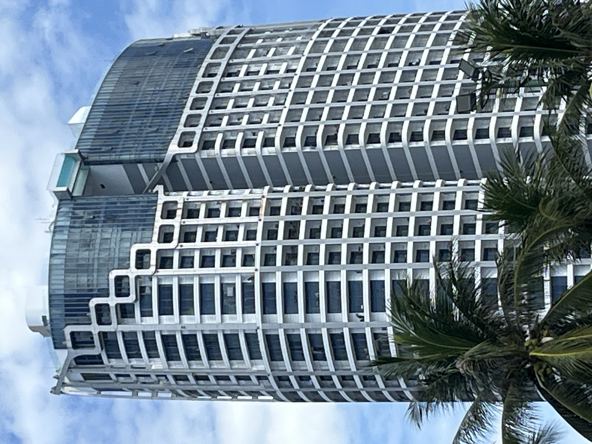 The image shows a tall, curved skyscraper in Nha Trang, Vietnam. The building has a modern design with a mix of white and gray facades, and large windows. The upper floors are clad in blue glass, and a rooftop pool is visible. Palm trees are in the foreground, obscuring the lower part of the building. The sky is blue with scattered white clouds. The overall impression is one of a luxury hotel or condominium complex.