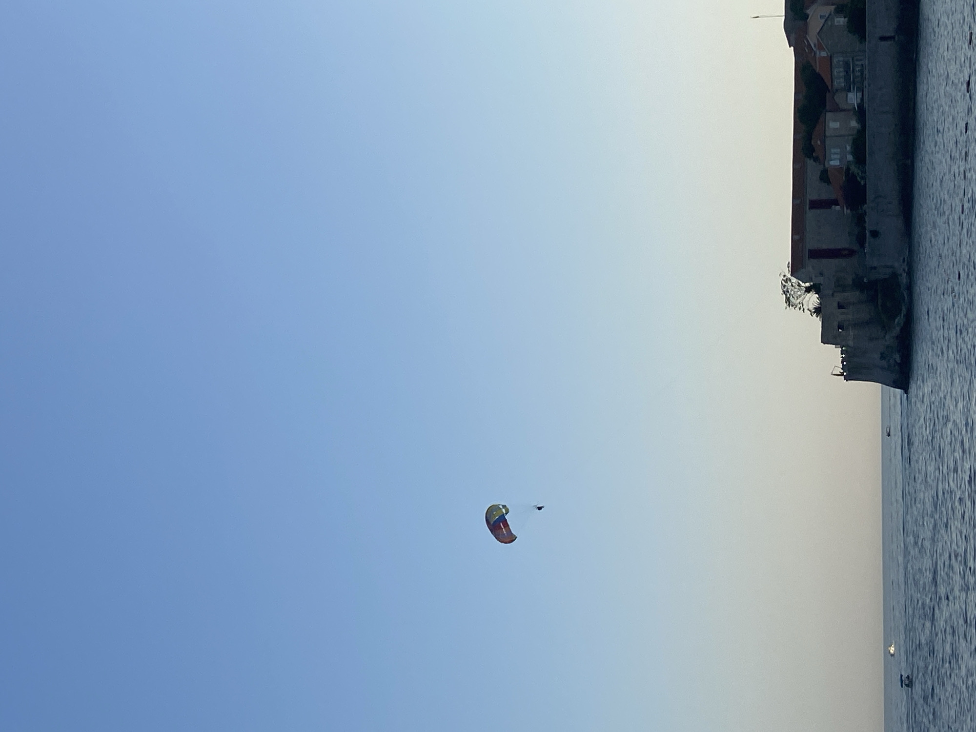 In the vast expanse of a clear blue sky, a brightly colored parachute soars, carrying a lone individual suspended beneath it. The parachute is a vibrant mix of red, yellow, green, and blue, creating a striking contrast against the serene sky. Below, the deep blue ocean stretches to the horizon, its surface gently rippled. On the right side of the image, a coastal town appears to be nestled on a rocky outcrop. The buildings are characterized by their red-tiled roofs and stone walls, suggesting a historical architectural style. Several boats are visible on the water, adding a touch of activity to the scene. The overall impression is one of a peaceful day with recreational activities taking place against a backdrop of natural beauty and historical architecture.