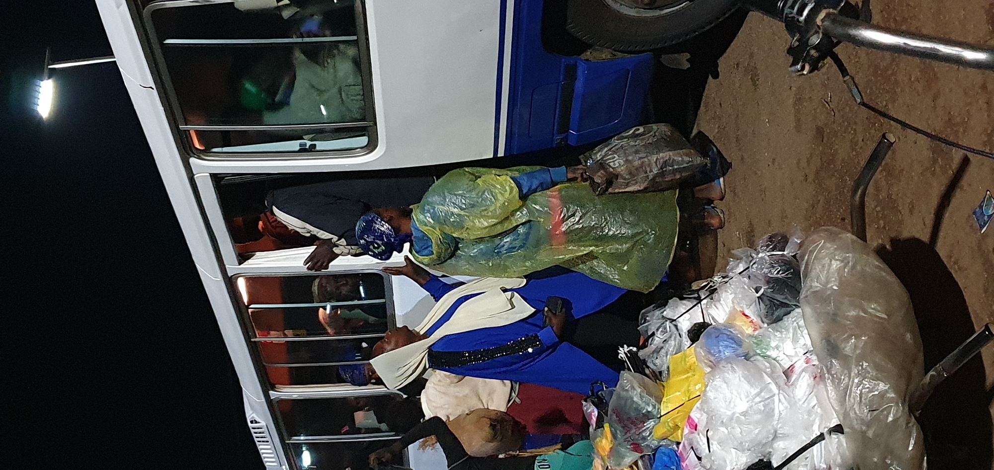 In Bukuru, Nigeria, at night, a group of people are gathered around the open door of a white and blue bus. One individual, dressed in a black robe with a white head covering, is standing in the doorway, appearing to interact with those outside. Several people are positioned near the bus entrance, some are wearing headscarves, including a woman in a blue and white outfit with bejeweled sleeves and another woman in a yellow translucent raincoat. In the foreground, a large pile of various plastic-wrapped items is present. The ground is dirt, and a bicycle handlebar is visible in the lower right corner. A street light illuminates the scene from above.