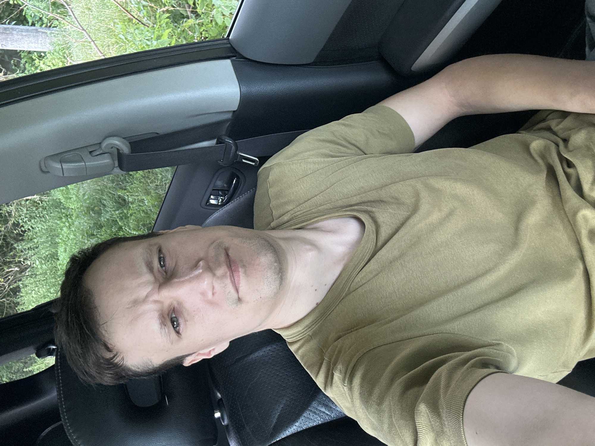 A man with short brown hair and a light complexion sits in the driver's seat of a car. He is wearing a tan, short-sleeved t-shirt. His expression is ambiguous, with furrowed brows and slightly pursed lips. The car's interior is visible, including a black leather seat, a gray seatbelt, and a door panel. Outside the car, lush green foliage and trees are visible, suggesting a rural or natural setting. The image does not provide information about the specific location within Makiv, Ukraine, beyond the general context of being in a vehicle.