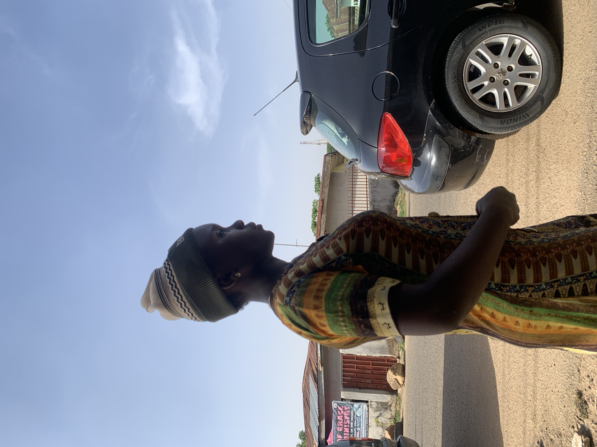A street-level, color photograph depicts a woman in Gura Top, Nigeria, standing on a paved road next to a parked, dark-colored vehicle. She is wearing a patterned, multicolored garment with stripes and a matching head covering. The woman is positioned in the lower center of the frame, her face turned upwards towards the clear, blue sky.
Behind the woman, a black car occupies the top right corner of the image, showing the rear tire, taillight, and a portion of the hatchback. Adjacent to the road, there are buildings and structures, including a church with a sign bearing the text "F GRACE MINISTRY" and "WP16 WINDA 205/65R15". The environment appears to be a residential or mixed-use area.