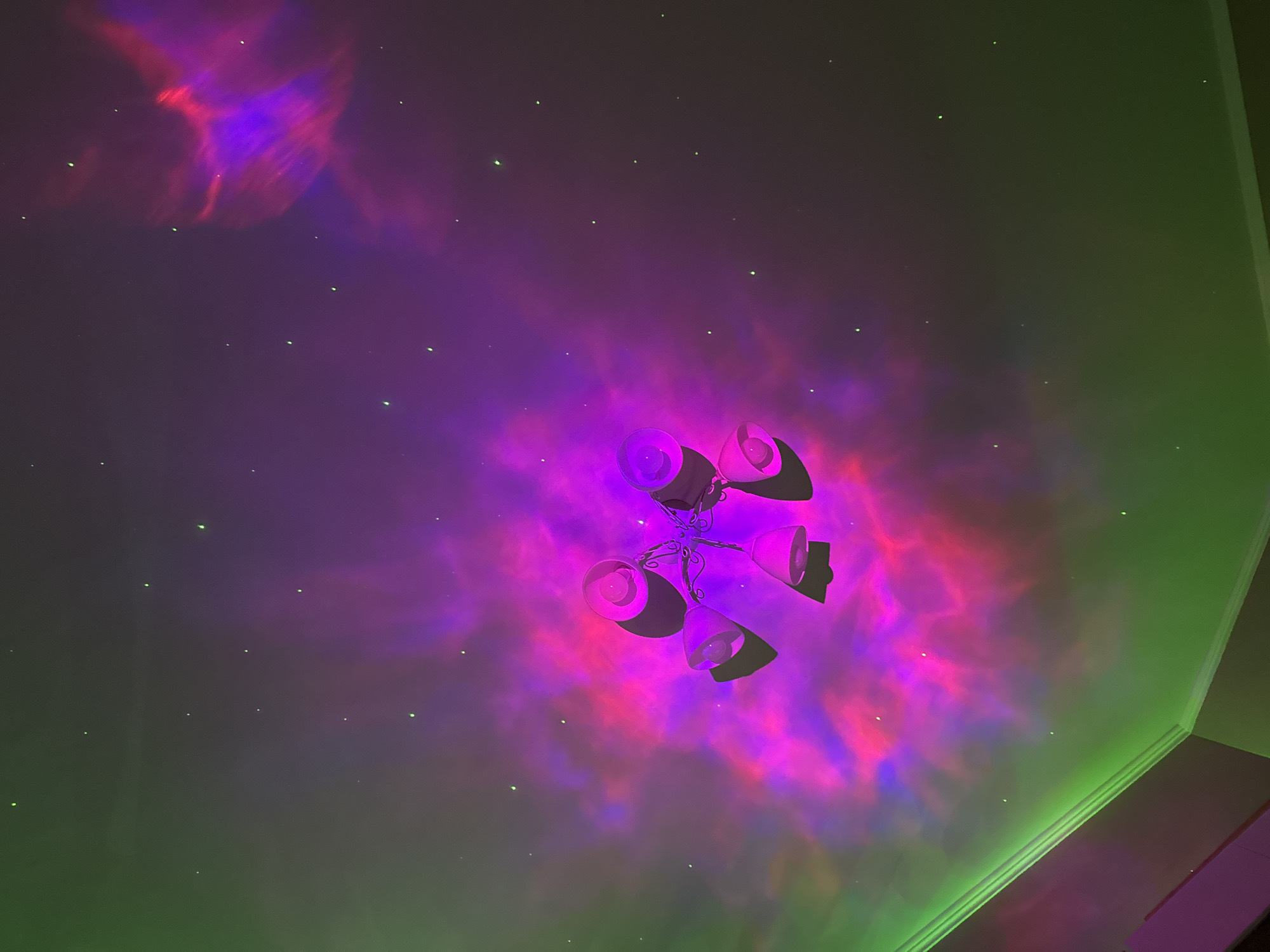 The image shows a ceiling illuminated by a projector creating a nebula effect with swirling purple and pink lights, and scattered green pinpoints resembling stars. A ceiling fan with four white lamps is positioned in the upper right quadrant of the image, casting shadows onto the colorful ceiling. The lighting creates a whimsical and cosmic atmosphere, suggesting a decorative lighting setup rather than a natural phenomenon. There are no people visible. The scene is dimly lit apart from the projector's output. The room's corner is visible on the right edge of the frame, with a plain wall extending upwards. The overall impression is one of a stylized, otherworldly environment.