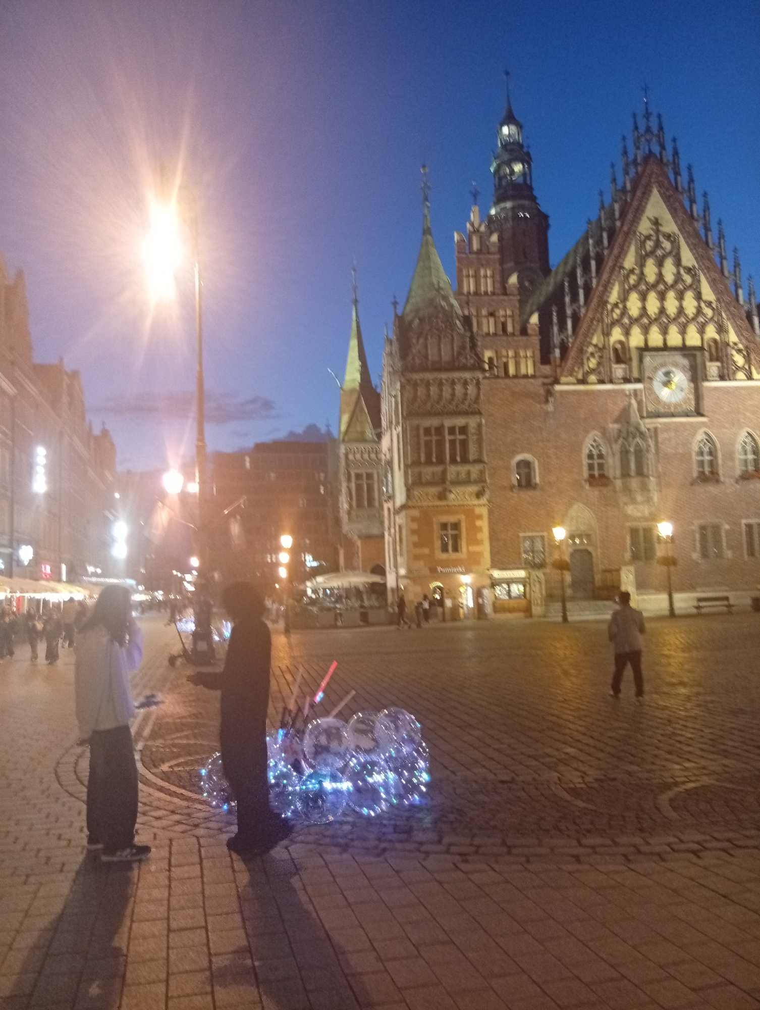 The image depicts a view of the Market Square in Wrocław, Poland, at dusk. The historic Old Town Hall is visible in the background with its characteristic Gothic architecture, adorned with decorative elements and a clock face. A street lamp with a bright light source is prominently positioned in the mid-ground, casting a warm glow. In the foreground, two individuals are standing on the cobblestone square. One person, dressed in lighter clothing, is facing away from the camera and appears to be interacting with the second person, who is facing the camera and wearing darker attire. Next to the second person, a collection of illuminated, transparent balloons is displayed. The surrounding area shows other buildings with illuminated windows and some people walking in the distance, suggesting a public space with some activity. The sky is a deep blue, transitioning from daylight to night.