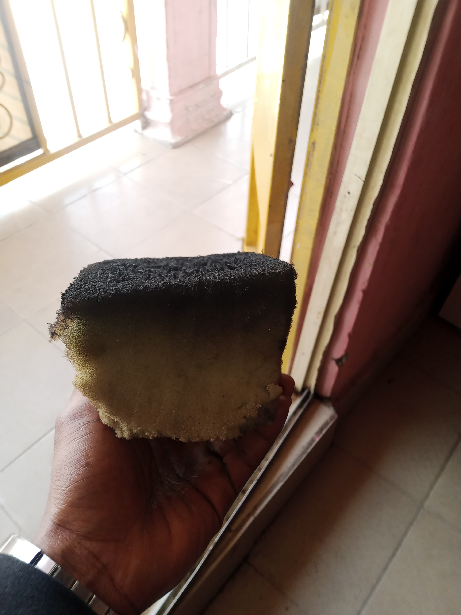 A person's hand, wearing a silver watch on the wrist, is holding a piece of burnt sponge. The sponge is dark and charred on one side, transitioning to a lighter color towards the bottom. The background appears to be an outdoor setting, possibly near a building with painted columns and a door or gate. The floor is tiled. The scene is illuminated by natural light. This image was taken in Jalingo, Nigeria.