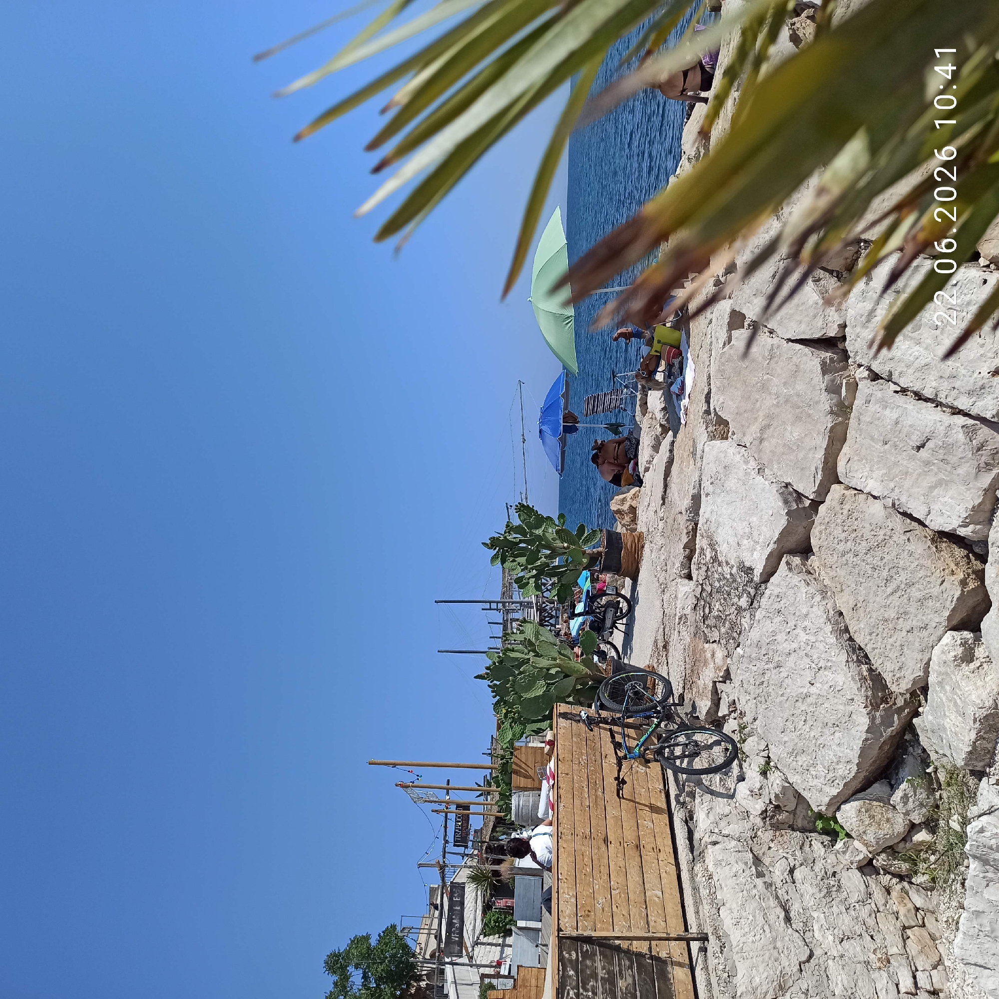 The image depicts a sunny day at a rocky beach with a view of the sea. In the foreground, large, light-colored rocks form the uneven terrain. To the left, a wooden structure with a slatted front is visible, and a bicycle is parked against it. Further back, a second bicycle is also visible. Several large potted cactus plants are strategically placed. In the middle ground, several people are relaxing on the beach, some under umbrellas, while others are sitting or lying on towels. A blue and a green umbrella provide shade. The sea is a deep blue, with gentle waves. In the background, there are some structures that appear to be part of a seaside establishment, including what looks like a fishing platform or pier with a net. The sky is a clear, bright blue. The date stamp "22.06.2026 10:41" is visible in the bottom right corner. Based on the context and the presence of a structure resembling a "trabucco," this scene is likely located in Trani, Italy, a coastal city known for such traditional fishing platforms.