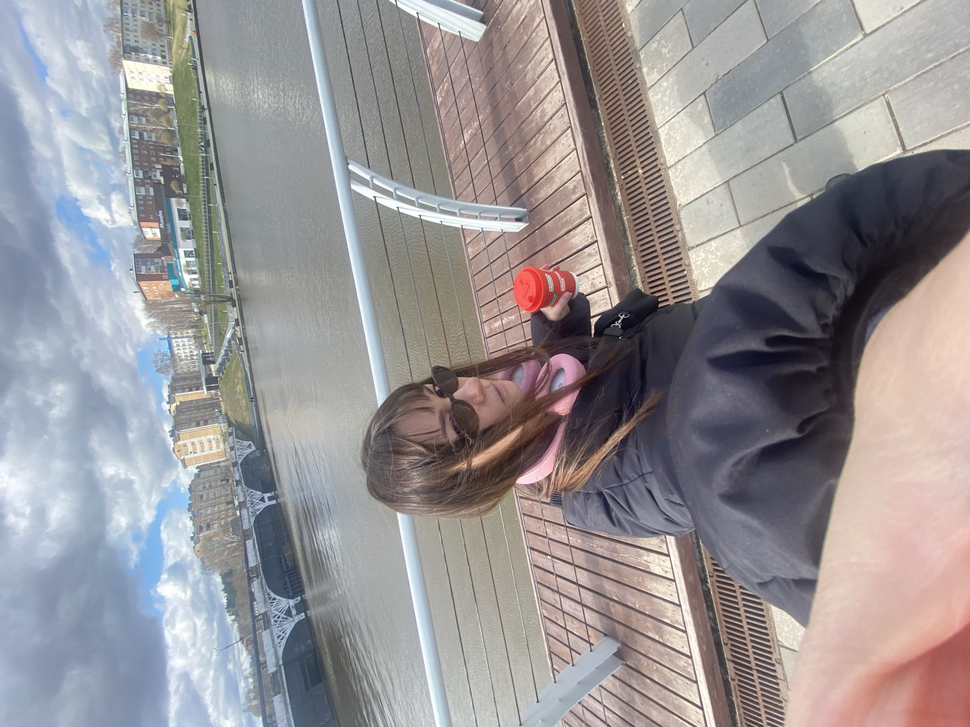 A young woman stands on a wooden deck next to a river in Votkinsk, Russia. The river is on her left and across the river are buildings. The woman is wearing sunglasses, a black jacket, and pink headphones around her neck. She is holding a red cup. To her right is a grey paved area with a drain. Behind her are white railings. The sky is cloudy.