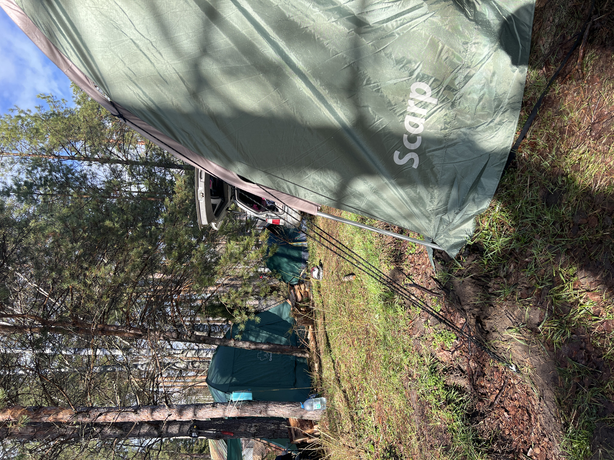 The image shows a forest campsite in Lesnoye, Russia, under bright daylight. A large olive-green tarp or tent canopy dominates the foreground and right side of the frame, featuring the white brand name "Scarp." This main shelter is secured by visible tent poles and dark guy lines extending to the ground, which consists of damp earth, sparse grass, and fallen pine needles.
In the mid-ground and background, several smaller green tents are pitched amidst numerous tall pine trees. One of these tents has a partial label, "Green," visible on its side. A light-colored car, possibly white or silver, is parked nearby with its trunk open, suggesting its use in the camping activity. The sky, visible through the tree canopy, is blue with some scattered white clouds, and sunlight casts distinct shadows, indicating a clear day. Various camping items, such as a plastic water bottle and what appears to be stacked firewood, are visible around the campsite. The ground shows areas of mud and uneven terrain, characteristic of a natural outdoor setting.
