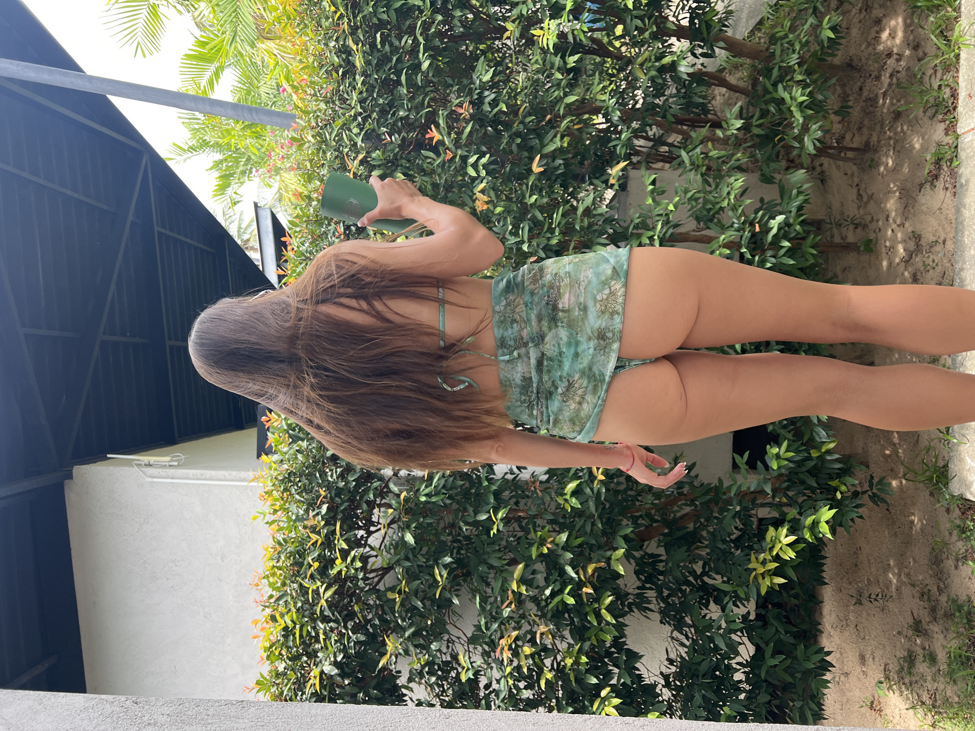 A woman stands with her back to the camera, wearing a teal bikini and a matching sarong. She is holding a green cup in her right hand. The woman has long, dark brown hair. She is standing in an outdoor area with lush green foliage behind her, and a white wall to her left. The ground is sandy with some sparse green grass. A dark roof structure is visible above the foliage. The scene appears to be in a tropical or subtropical climate, possibly a resort or a private residence in a location like Ban Bang Rak, Thailand.