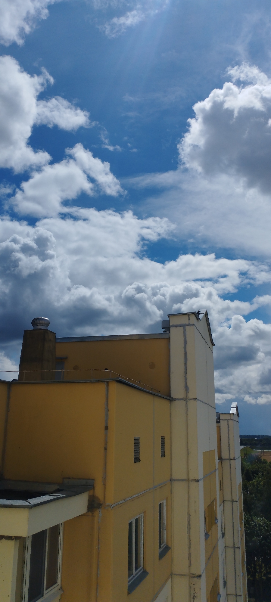 The image is a vertical, eye-level shot of a multi-story building's upper facade against a partially cloudy blue sky. The building is primarily painted a bright yellow, with cream-colored accents and vertical structural lines. A prominent, dark grey chimney with a silver vent cap is visible on the left side of the building. Several windows, some with white frames and others with dark frames, are present on the yellow sections, along with a few narrow, louvered openings. The cream-colored sections feature decorative, stepped architectural elements at the top. Two birds are perched on the roofline of the cream-colored section on the right. In the distance, a hazy landscape with trees and a road is visible. The sun is casting a bright glare through the sky on the upper right portion of the image.
