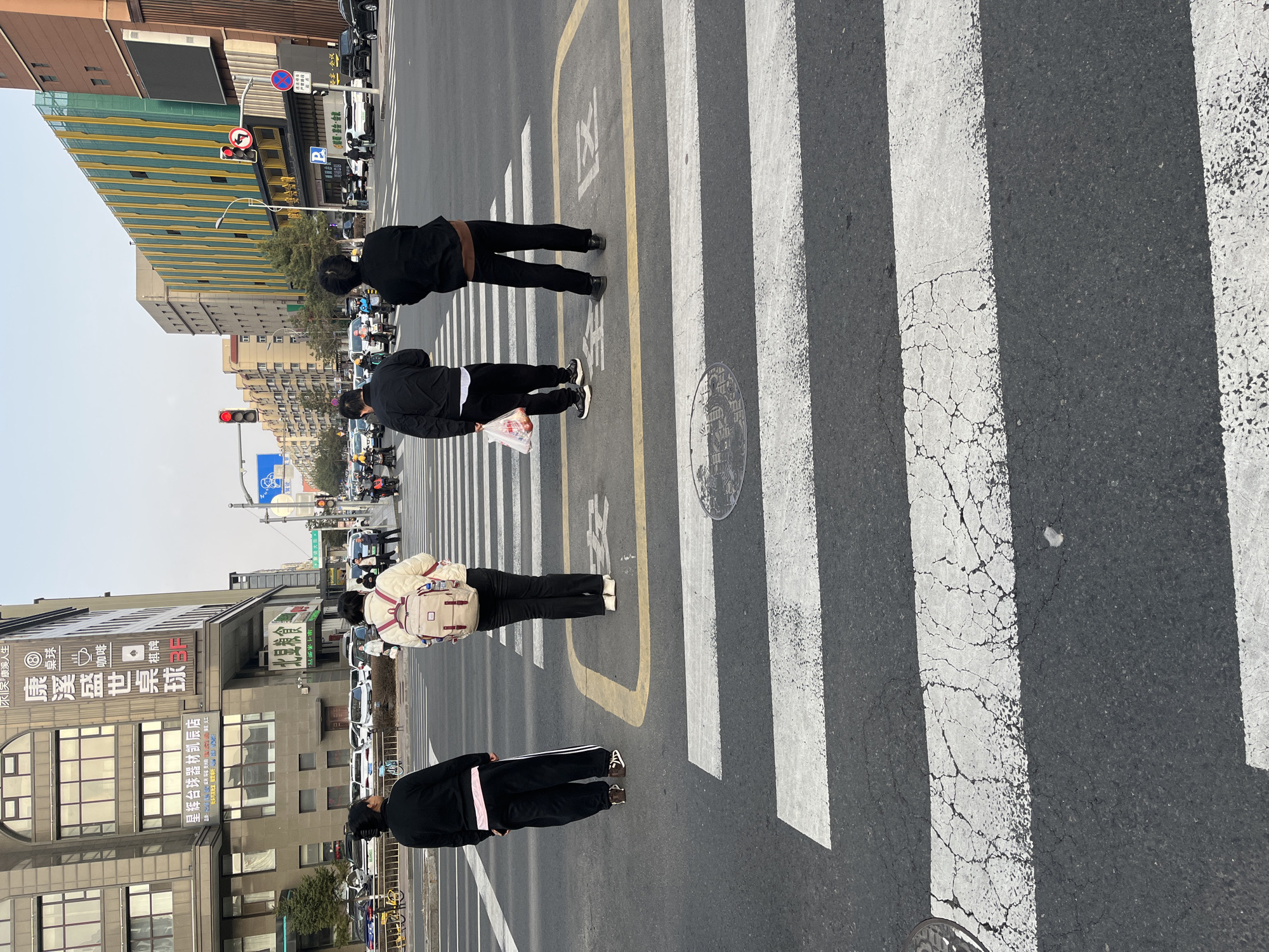 This media file captures an urban street scene in Changchun City, China, likely at an intersection with a crosswalk. The image is oriented vertically but depicts a horizontal view across the street.
Four individuals are visible from behind, standing on or near a pedestrian waiting area marked with yellow lines and the Chinese characters "待区" (waiting area) on the asphalt. They appear to be waiting to cross the street. The person closest to the viewer wears a black tracksuit with white stripes. To their right, another individual wears a light-colored puffer jacket and a matching backpack. Further right, a person in a black jacket and black pants holds a white plastic bag. The fourth person, also in a black jacket and pants, stands on the far side of the crosswalk.
The road features a clearly marked pedestrian crosswalk with white stripes, some showing cracks and wear. A manhole cover is also visible on the crosswalk. In the background, several multi-story buildings line the street. One prominent building exhibits a striking yellow and green striped facade. Other buildings are more subdued in color, one displaying signage for "康溪盛世桌球" (Kangxi Shengshi Billiards) and "星辉台球器材凯辰店" (Xinghui Billiards Equipment Kaichen Store), along with "桌球" (Billiards), "咖啡" (Coffee), and "棋牌" (Chess and Cards). Another sign mentions "水天康溪人生" (Shui Tian Kang Xi Ren Sheng).
Multiple traffic lights are visible, with red lights illuminated, suggesting that vehicles are stopped and pedestrians are awaiting their turn to cross. Various road signs are present, including a blue sign with a bicycle symbol, a red circular 'no stopping' or 'no parking' sign, a 'no left turn' sign, and a blue 'P' (parking) sign. Cars and motorcycles are parked or waiting along the street. The sky appears clear, and the scene is set during daytime, with diffused lighting. The overall impression is a bustling city environment.