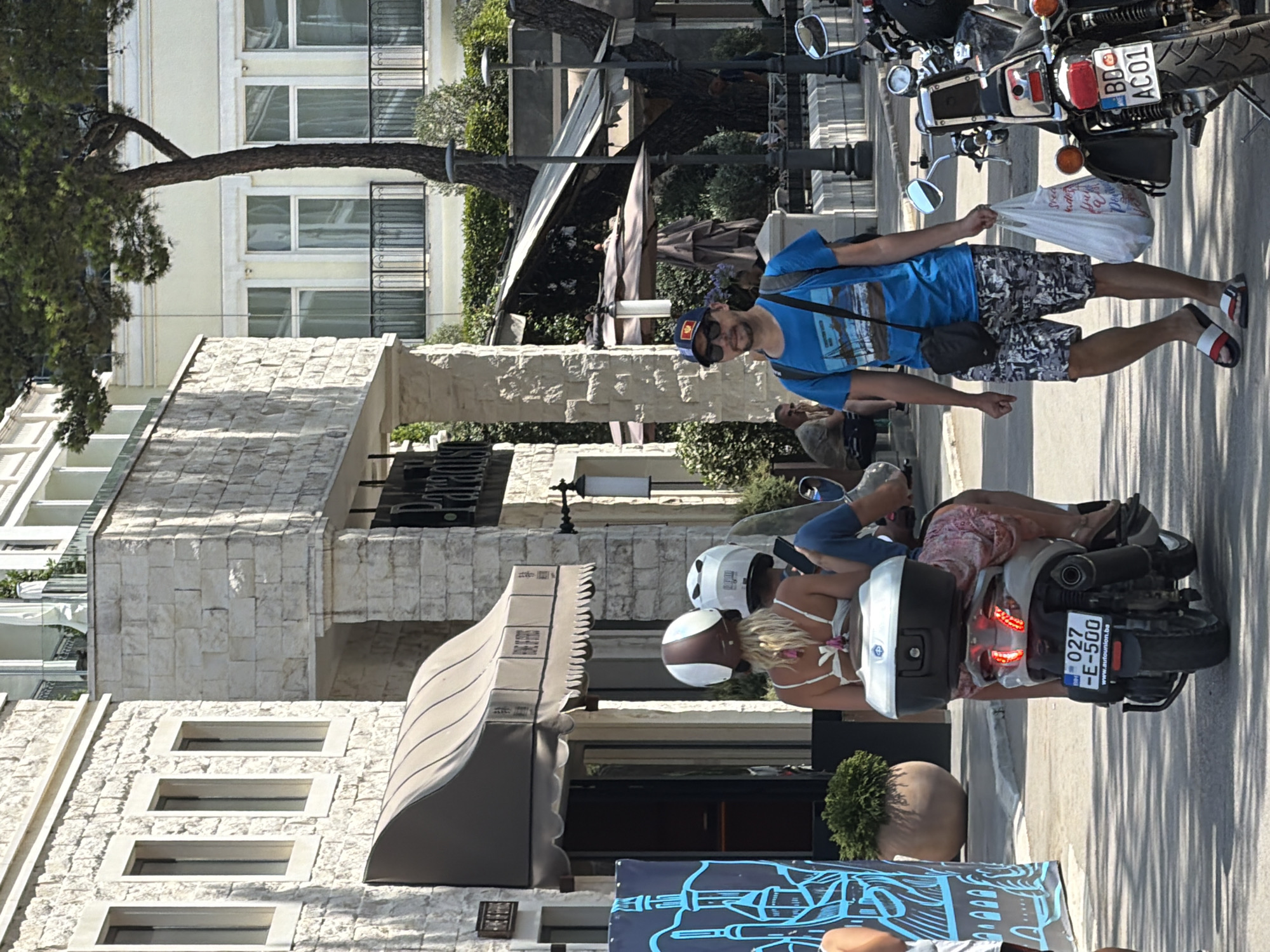 In Budva, Montenegro, on a sunlit street, a man wearing a blue t-shirt and patterned shorts walks towards the viewer. He carries a white plastic bag in his right hand and a black crossbody bag over his left shoulder. He is wearing a blue baseball cap with a Montenegrin flag patch and sunglasses. To his left, a scooter carries two individuals wearing helmets. The rider in front has blonde hair and wears a white bikini top, while the person behind wears a blue t-shirt and blue pants. The scooter has a silver storage box on the back and a license plate that reads "027 E-500." The background features a building with a stone facade and awnings, as well as trees and streetlights. To the right of the man, a parked motorcycle with a license plate reading "BD AC01" is visible.