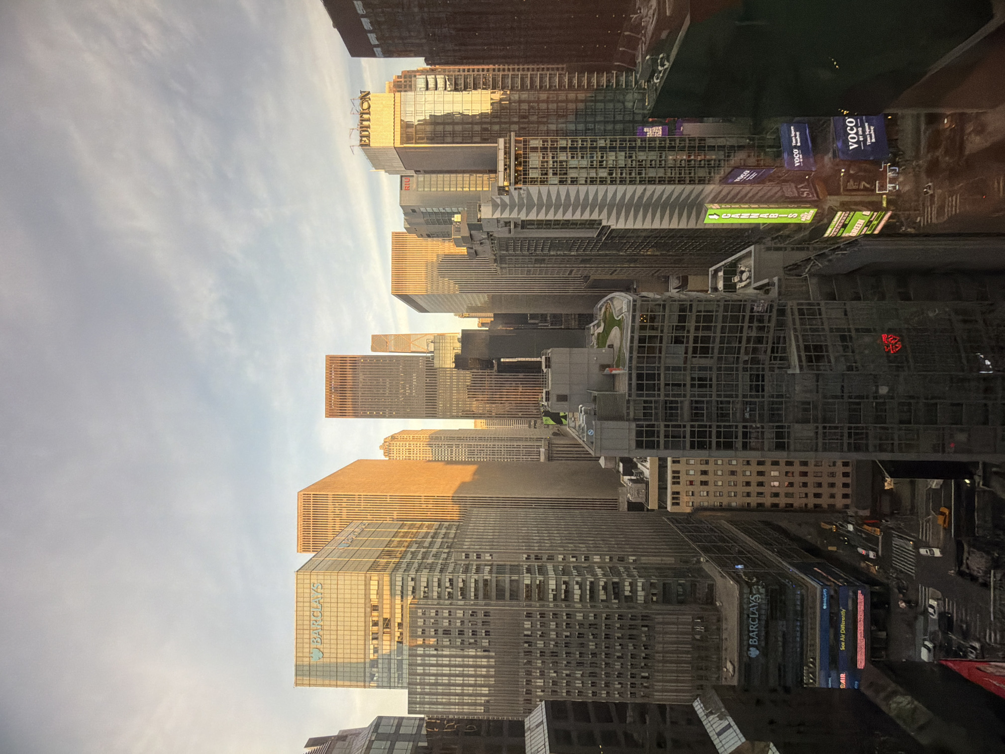 This media file captures a high-angle view of a dense urban cityscape, likely New York, USA, under the warm glow of what appears to be late afternoon or early evening. The scene is dominated by numerous skyscrapers of varying heights and architectural designs, some with reflective glass facades and others with more textured exteriors.
The time of day is notable for the "golden hour" lighting, where buildings on the left side of the frame are illuminated by soft, golden sunlight, casting long shadows and creating a stark contrast with buildings in shadow further into the city. The sky above is a pale blue with diffused, wispy clouds. No specific human activity or people are distinctly visible, but the presence of street-level signs and faint indications of traffic suggest a bustling metropolis.
Key visible text and brand names include:
* "EDITION" prominently displayed on a golden-hued building.
* "BARCLAYS" appears on several buildings and signs, one of which also reads "See Air Differently" and "DAIR."
* Multiple illuminated billboards feature "VOCO" and "BY IHG."
* Other signs display "CANNABIS," "GREEN," and "RIU."
The overall setting is that of a major commercial and financial hub, characterized by its towering architecture and vibrant illuminated signage.