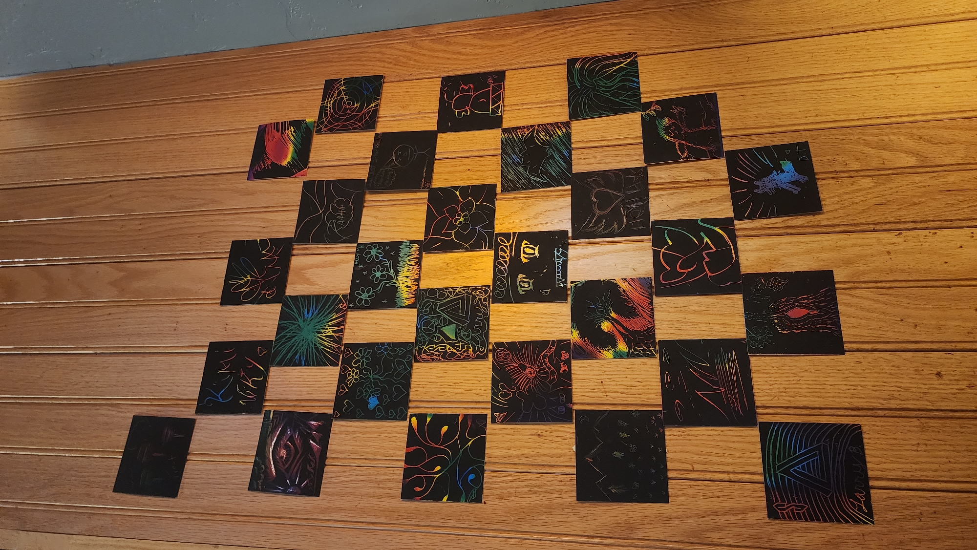 The image shows a collection of scratch art pieces arranged on a light brown wood-paneled wall. The wall is likely an interior wall, given the lack of exterior features. The scratch art pieces are small, square or rectangular, and feature various colorful designs revealed by scratching away the black surface. The designs include abstract patterns, floral motifs, faces, and other recognizable shapes. The composition of the arrangement is somewhat grid-like but not perfectly aligned. There is no indication that the image was taken in Jackson, United States of America. There are no people in the image.