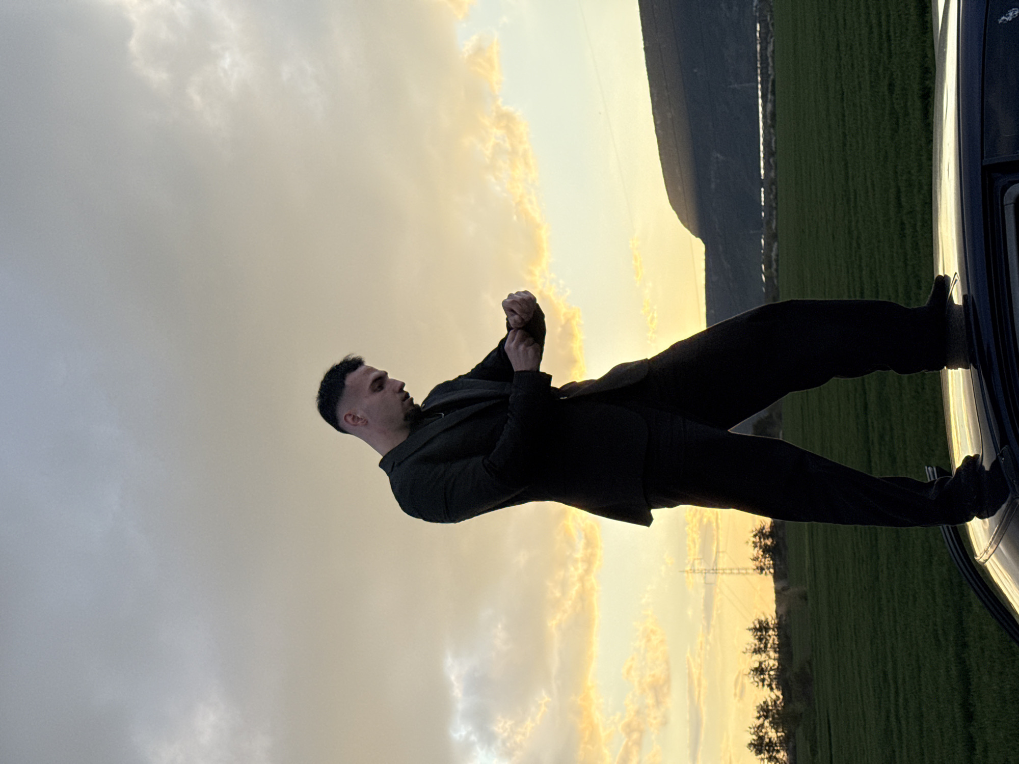 A man in a black suit stands in what appears to be a field. The sky is filled with clouds lit by the setting sun, casting a golden glow on the scene. To the right, a vehicle is parked, and behind it is a green field with mountains in the distance. Based on the surrounding landscape features like mountains and green fields, this could potentially be a location on the outskirts of Shumen, Bulgaria, perhaps near one of the rural areas surrounding the city.
