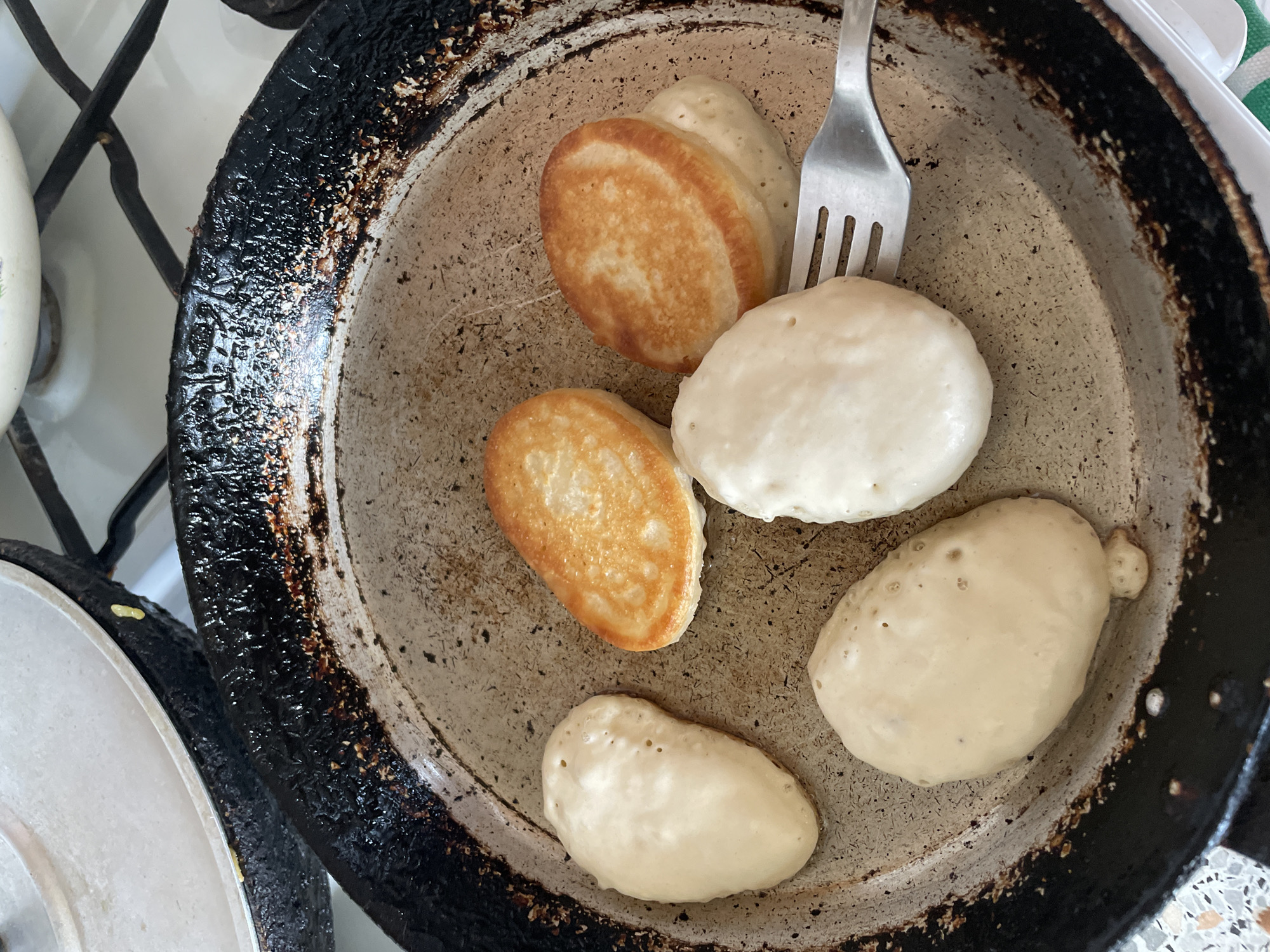 The image depicts a frying pan on a stovetop, likely indoors. The pan contains several pancakes in various stages of cooking, some golden brown and others pale. A metal fork is present in the pan, seemingly being used to maneuver the pancakes. The pan appears well-used with a blackened exterior. The setting is likely a residential kitchen. The city of Talmenka, Russia is only a location detail provided by the user and is not visible in the image.
