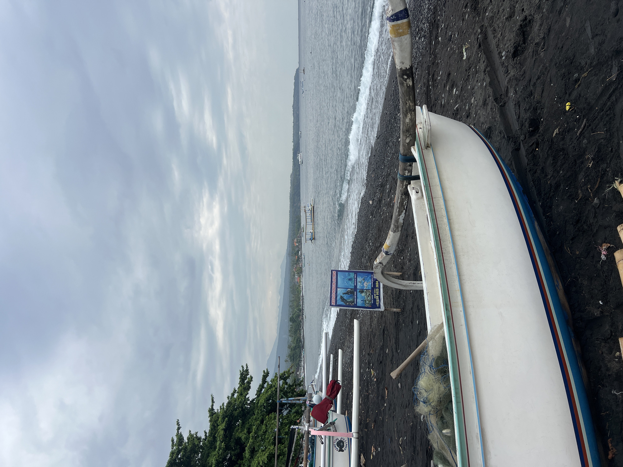 In Abang, Indonesia, a black sand beach is lined with traditional Balinese jukung boats. In the foreground, a white jukung with blue and red trim sits on the dark sand, its hull partially visible. A fishing net and a wooden oar rest inside the boat. Further down the beach, several more jukung boats are pulled ashore. A sign advertising "AHED BAGUS SNORKELING AND FISHING" stands near the water's edge. The sea is calm with gentle waves breaking on the shore. In the background, lush green trees line the coast, and mountains rise in the distance under a cloudy sky. Several boats are visible on the water, suggesting a moderate level of activity. The overall scene depicts a tranquil coastal village environment in Indonesia.
