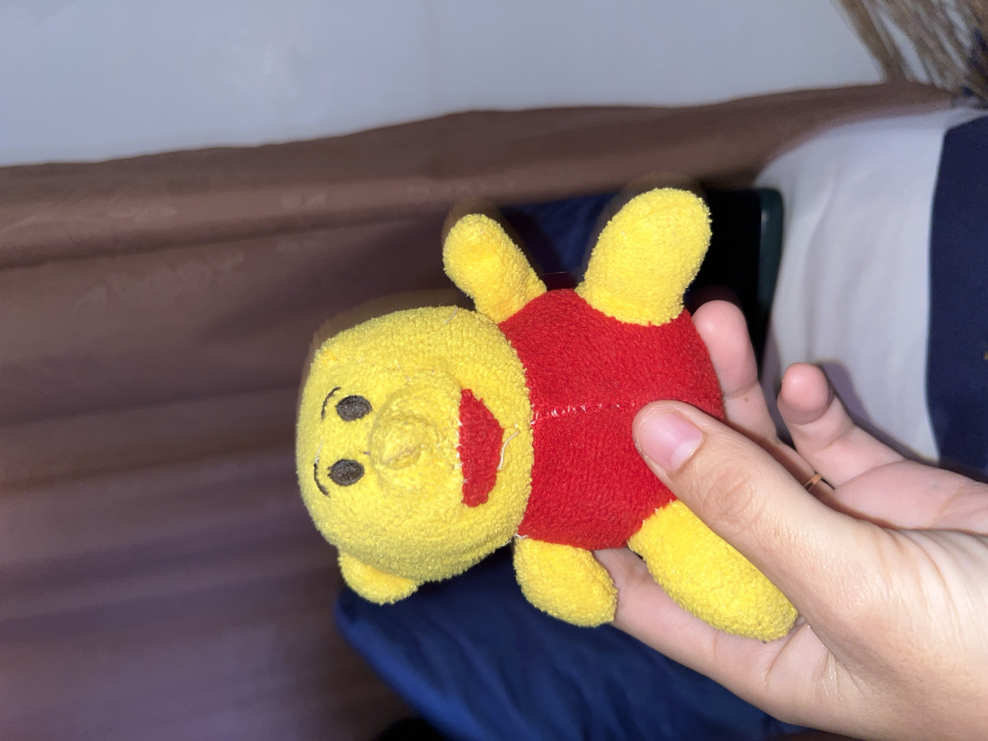A medium-close-up photograph captures a human hand holding a small, yellow and red plush toy. The toy, made of a soft, textured fabric, features a yellow head with simple stitched black oval eyes, curved eyebrows, a protruding round nose, and a red smiling mouth. Its torso is red, while its limbs are yellow. Loose threads are visible around the toy's mouth and where the arm attaches to the body. The hand holding the toy displays a light-to-medium skin tone, with the thumb, index, and middle fingers visibly gripping the lower portion of the toy. A thin, metallic ring is present on the ring finger. The background consists of various fabrics and surfaces: a brown, vertically folded fabric (likely a curtain) in the upper right, a plain white vertical surface, and dark blue fabric in the upper left and lower center. In the lower right, a white fabric with a horizontal dark blue stripe, possibly part of clothing, is visible, along with a lighter brown woven texture. The scene is brightly illuminated, indicating an indoor environment.