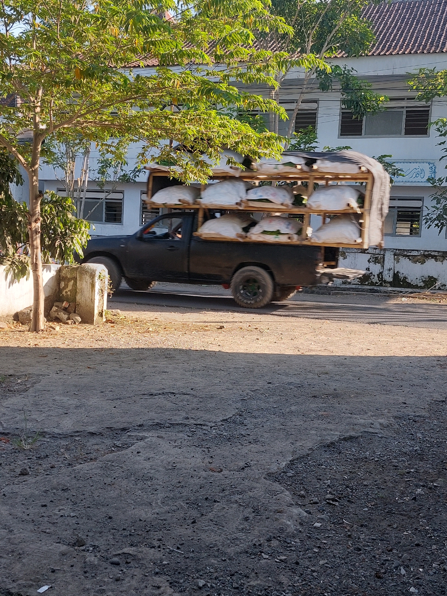 The image captures a black pickup truck driving on a street in Ajung, Indonesia. The truck is heavily loaded with white sacks stacked on a wooden rack in its bed. The truck's tires are blurred, indicating motion. The truck is passing by a white building with a tiled roof, and a tree with green foliage is visible on the left side of the frame. The foreground shows a dirt road with some gravel and debris. The lighting suggests it is daytime, possibly late afternoon given the shadows.
