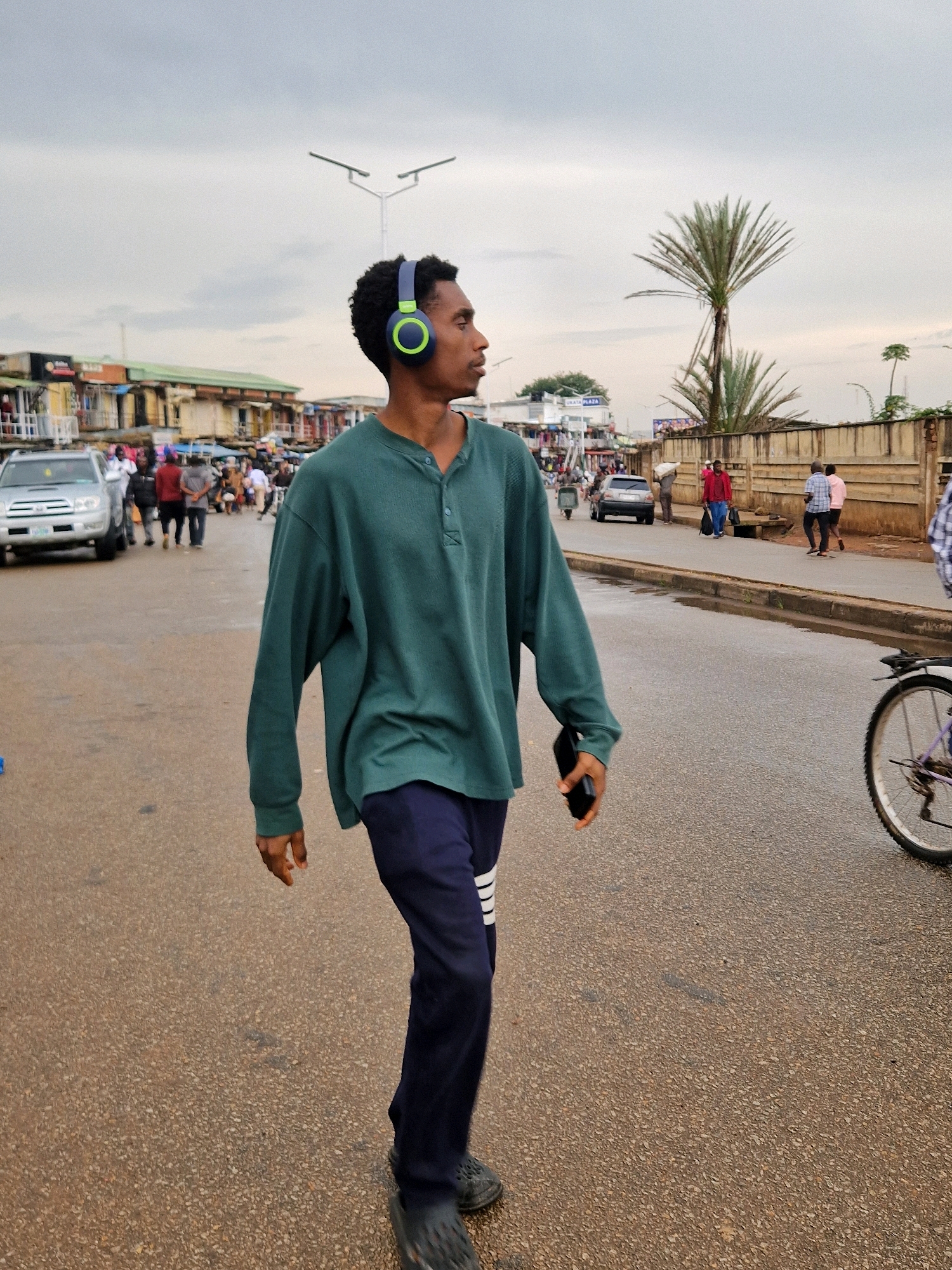 A young Black male walks on a wet, asphalt road in an urban area. He is wearing a teal long-sleeved henley shirt, dark blue sweatpants with white stripes on the sides, and black clogs. He has headphones on, which are teal and green, and is holding a black smartphone in his right hand. The man is walking away from the camera and towards the right side of the frame. In the background, there are buildings, palm trees, streetlights, cars, and other pedestrians. The sky is overcast.