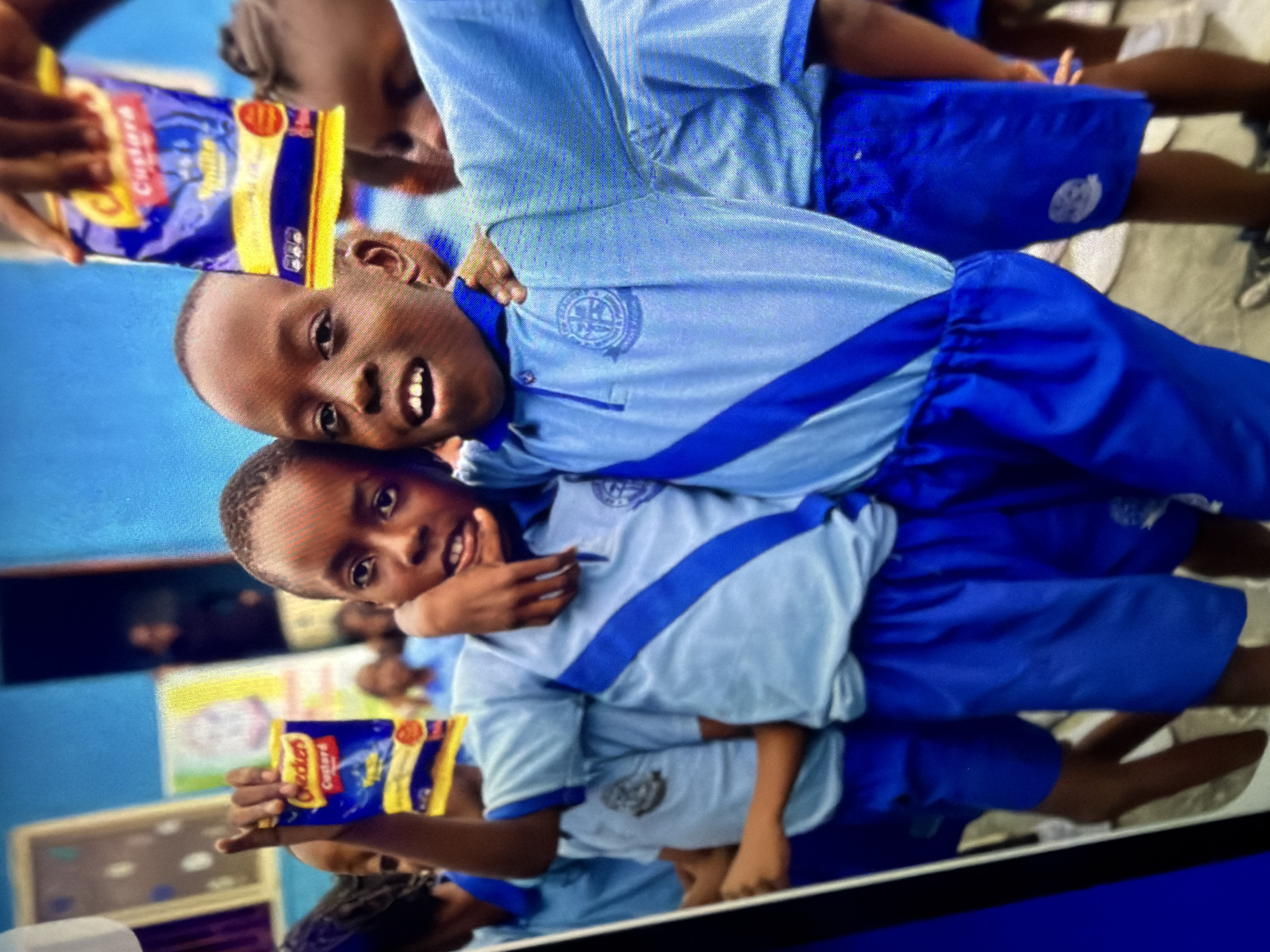 In this close-up, medium shot photograph, two young boys, likely students, are the central focus. They are dressed in matching light blue school uniforms with darker blue accents on the collars and diagonal stripes across their chests. The boy on the left has his arm around the boy on the right, and both are looking towards the camera. The boy on the right is holding up a small, blue and yellow packet of "Checkers Custard" with "Vanilla" written on it, showing it to the camera. They appear to be in an outdoor setting, possibly at a school or community event, with a bright blue wall visible in the background and hints of other children and structures. The overall scene suggests a cheerful and positive moment. The location is Unknown, Unknown.