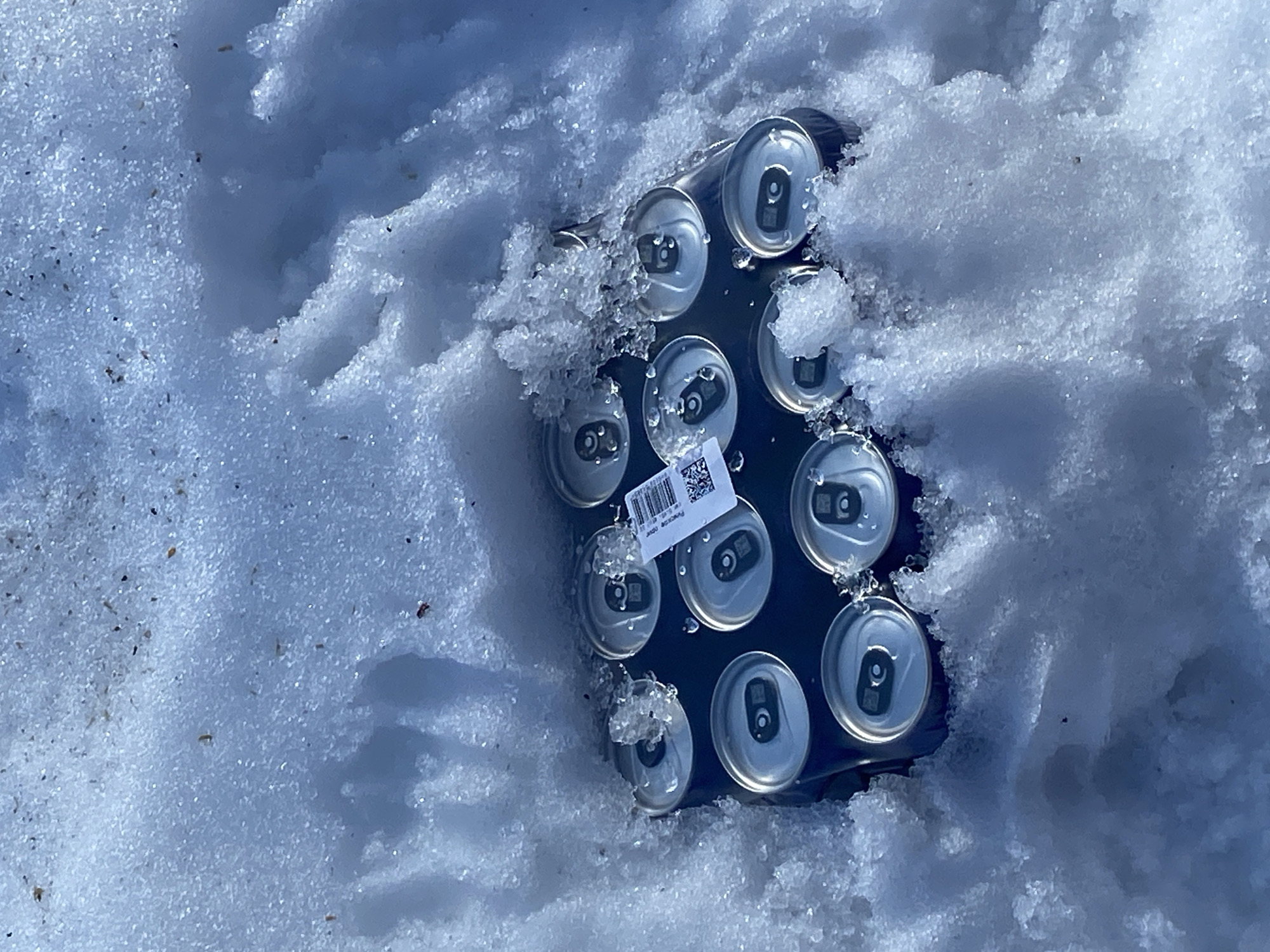In the city of Noviy, Russia, a dark-colored 12-pack container holding twelve silver-topped beverage cans is partially embedded in a field of white, granular snow. The cans are arranged in a grid, with their silver pull-tab mechanisms visible. A white adhesive label, approximately 5 cm x 2.5 cm, featuring a barcode, a QR code, and partially legible text including "PACKING DOSE," is affixed to the top of one central can. Small ice crystals and particles of organic debris are visible on the snow and around the can tops. The scene is illuminated by natural daylight, and no individuals or active human actions are depicted.