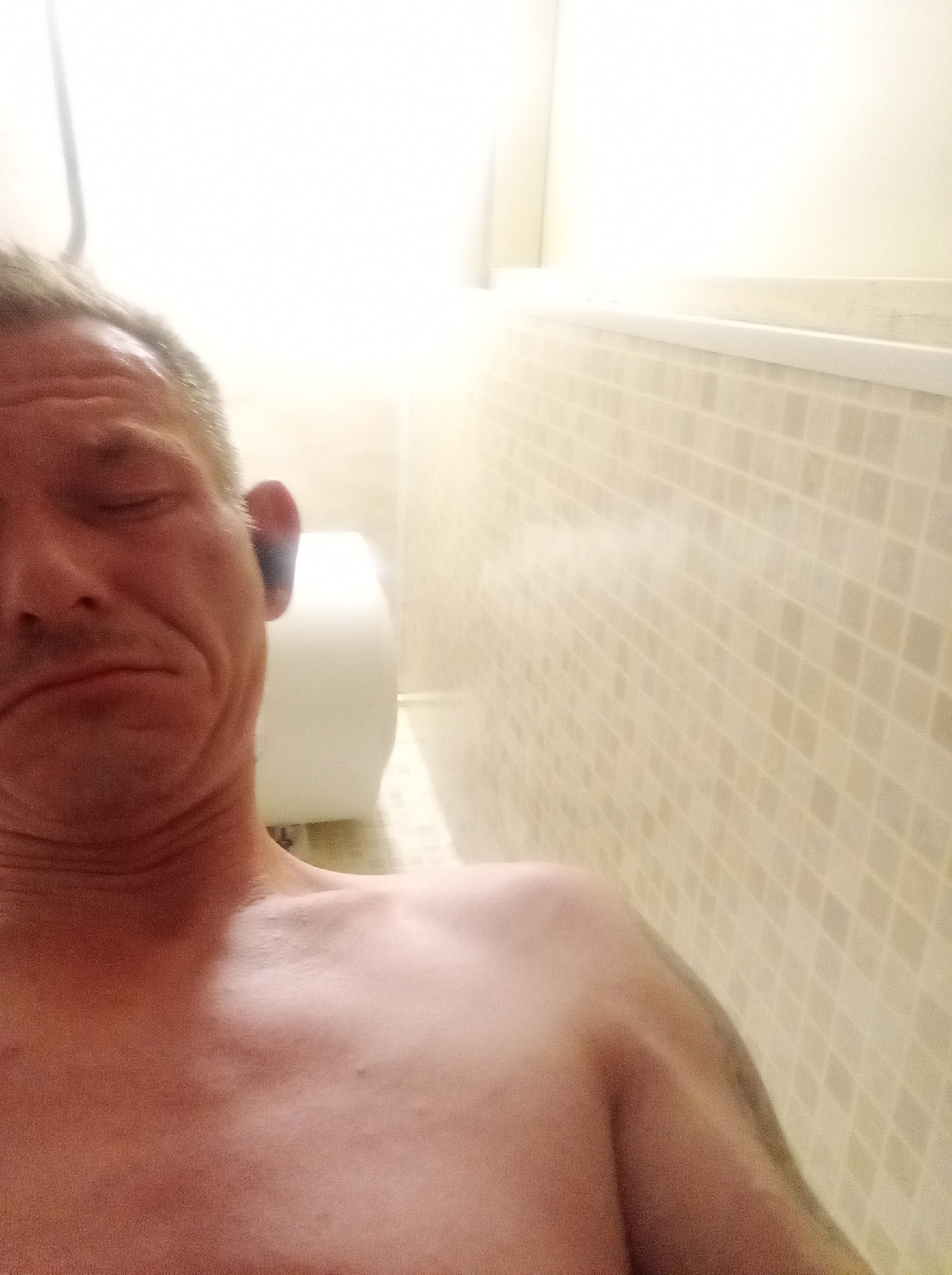 A man's upper body and head are pictured in a bathroom. He is looking at the camera with his eyes closed and mouth in a pucker. The background is a tiled wall, a toilet, and a bright light source. The man appears to be in a shower or bathtub.