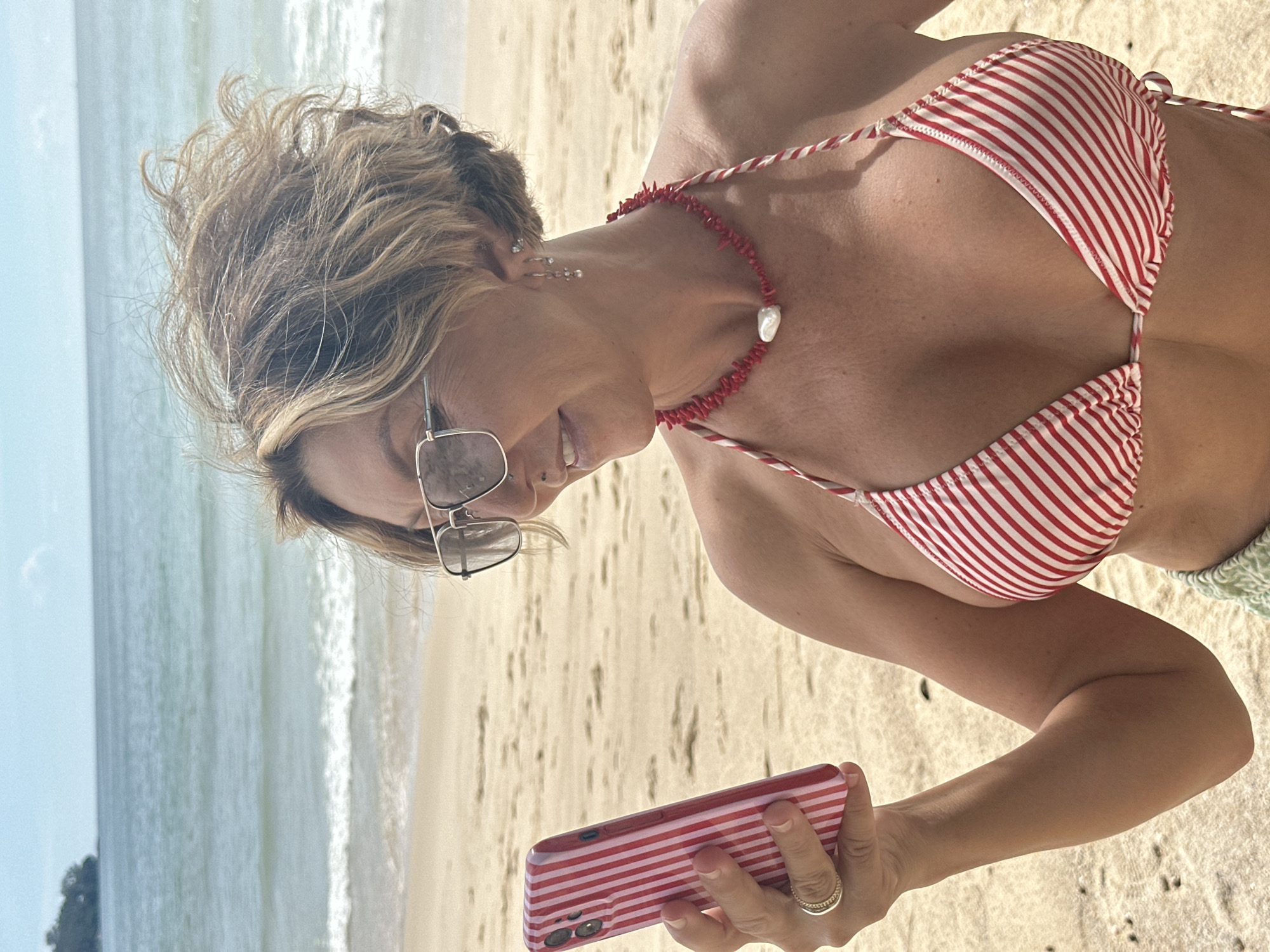 A woman with short, wavy blonde hair and sunglasses is on a sandy beach, holding a pink and white striped smartphone. She is wearing a red and white striped bikini top and a red coral necklace with a pearl. The ocean and sky are visible in the background. The woman appears to be looking at her phone.