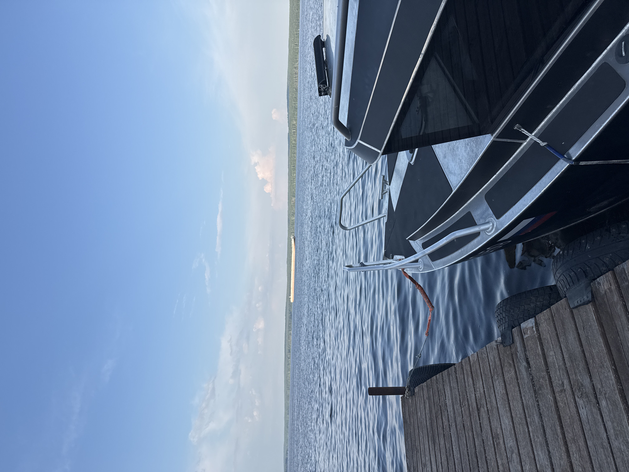 The image depicts a boat docked at a wooden pier on a calm body of water, likely the Kama River, near Chaikovsky, Russia. The boat, which is black with silver trim and a Russian flag emblem, is tied to the pier with a red rope. The pier is made of weathered wooden planks and has tires attached as fenders. In the distance, a large barge is visible on the water, with a tree-lined shore behind it. The sky is partly cloudy with blue patches. The perspective is from the boat looking towards the pier and the water.