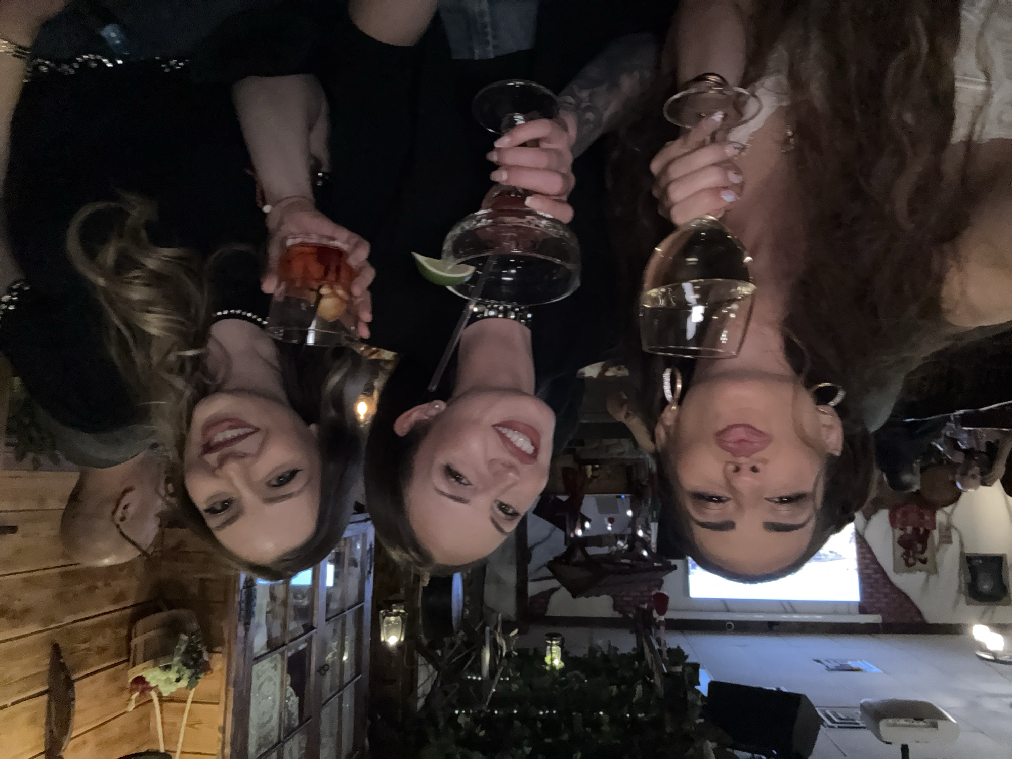 Three women are holding up glasses of different drinks in what appears to be a restaurant setting. The woman on the left is holding a small glass of amber liquid; the woman in the middle is holding a margarita; and the woman on the right is holding a glass of white wine. The restaurant has a rustic interior, with wooden walls and vintage-style lighting fixtures. In the background, there's a brick wall and what looks like a projector screen. The location is identified as Des Plaines, United States of America.