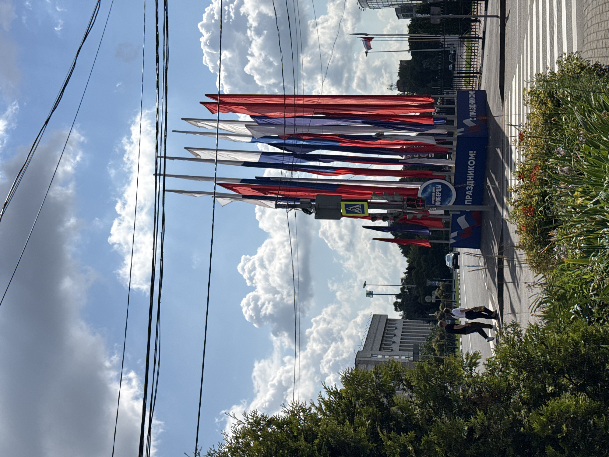 The image depicts a street scene in Tomilino, Russia, on a partly cloudy day. A prominent display of Russian flags is set up, featuring multiple red, white, and blue flags on poles. In the foreground, there is a lush green tree on the left and a flowerbed with various plants, including red flowers, in the lower right corner. A pedestrian crossing is visible in the middle ground. Two individuals are walking across the street at the pedestrian crossing. Behind the flags, a blue banner with white text, reading "С ПРАЗДНИКОМ!" (Happy Holiday!), is displayed. Further in the background, a building with a light-colored facade and rows of windows is visible on the left. A street light is also present. In the far background, trees and other buildings, including a tall cylindrical structure, can be seen. Power lines stretch across the upper portion of the image. A car is driving on the road in the middle ground.