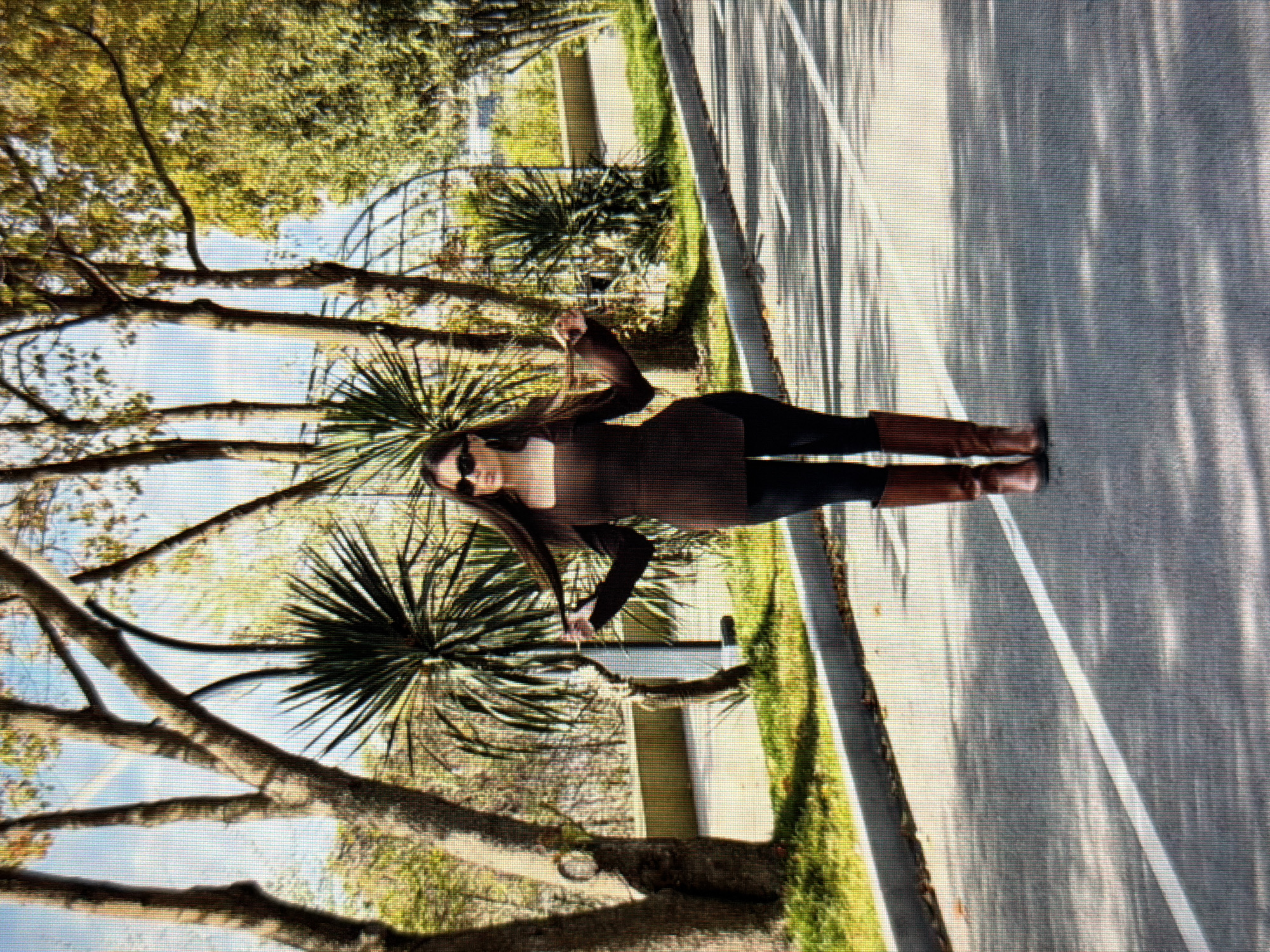 The image shows a woman standing on a paved road, possibly in a park or along a tree-lined street. She is wearing a brown top, black pants, and brown boots. She is holding her hair in both hands, with her arms extended to the sides. The background features trees, grass, and a clear sky. The scene is illuminated by sunlight, casting shadows on the road. There are no discernible landmarks or specific details that would place this image in Sirius, Russia.