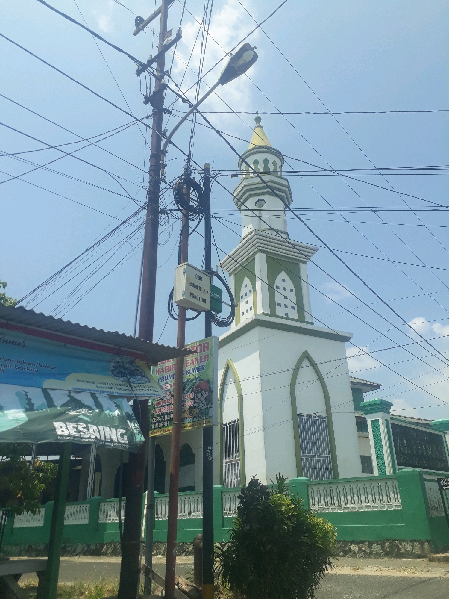 The image shows a white mosque with a green-and-white striped minaret, topped with a golden dome, against a bright blue sky filled with scattered clouds. Power lines crisscross the sky, with a utility pole in the foreground. To the left of the mosque, a green awning with text is visible, and a green fence runs along the front of the property. The mosque appears to be located in a built-up area, with glimpses of buildings and trees in the background. There are no people visible in the image. The overall scene suggests a sunny day in an urban or semi-urban Indonesian setting. The presence of the mosque minaret and the Islamic calligraphy on the awning indicate a location in a predominantly Muslim area, likely in Indonesia.