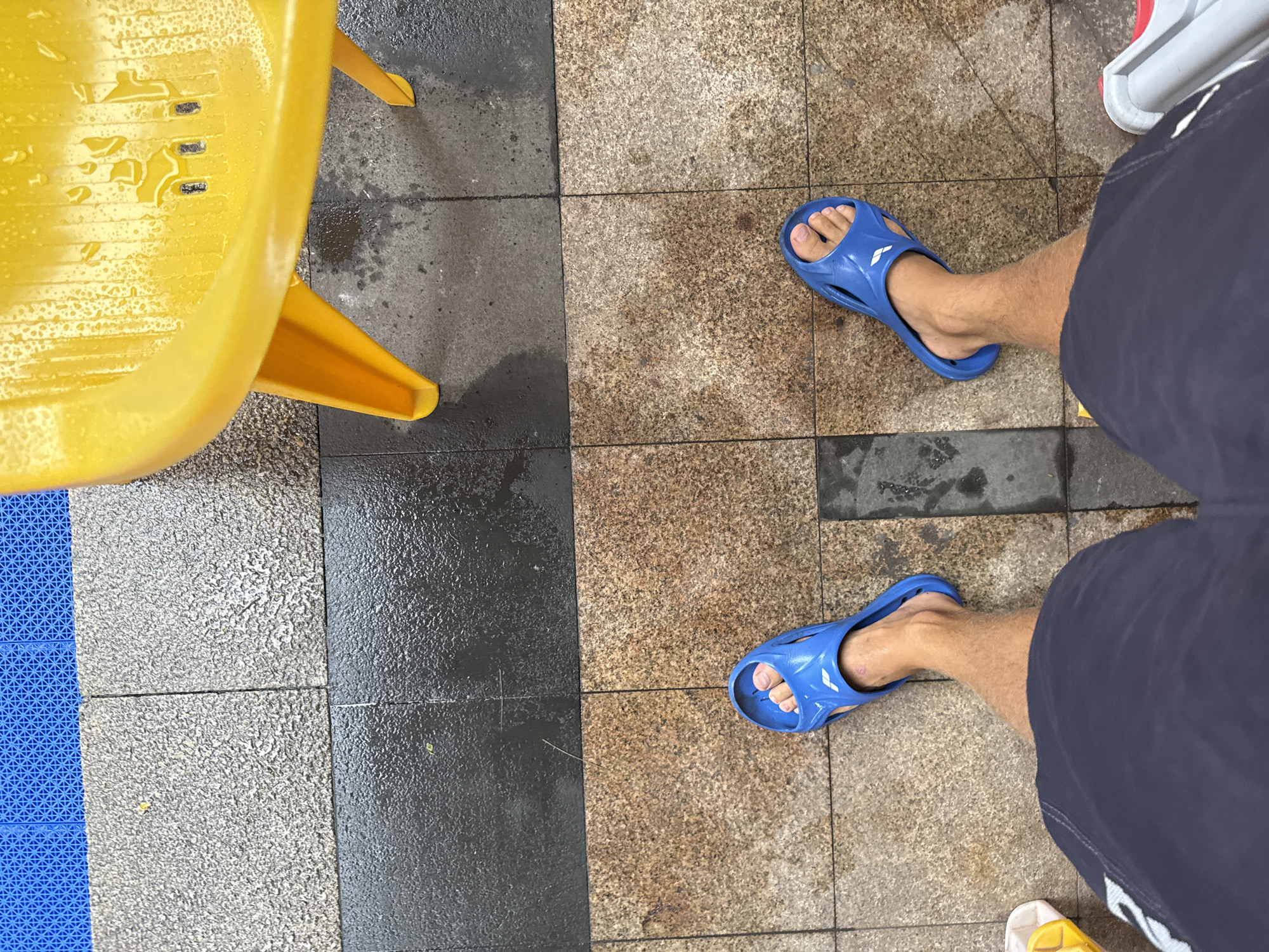 A first-person perspective shot shows a person's legs and feet from a slightly elevated angle. The person is wearing dark blue shorts and bright blue slide sandals. The sandals have a white logo on the straps. The ground is made of square tiles, with a section of speckled brown tiles and a section of dark grey, wet tiles. To the upper right of the frame, a bright yellow plastic chair is visible, also wet with water droplets. The background shows a section of blue interlocking mats. The scene appears to be outdoors, possibly after rain. The location is in Haizhu District, China.