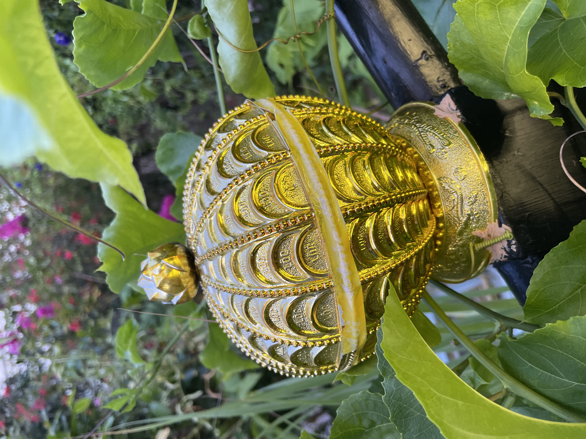 A golden, ornate sphere-shaped object, possibly a finial or decorative element, rests on a dark, cylindrical surface, likely a railing or support. The object is positioned outdoors, amidst lush green foliage, suggesting a garden or natural setting. In the background, a bokeh effect blurs vibrant pink and purple flowers, adding a pop of color to the scene. The golden sphere itself is intricately detailed, with raised, scale-like patterns and a series of smaller, bead-like embellishments along its circumference. A thin, translucent yellow band encircles the middle of the sphere. The overall impression is one of decorative opulence set against a natural backdrop. This image does not contain information to determine its location in Phan Thiet Ward, Vietnam.